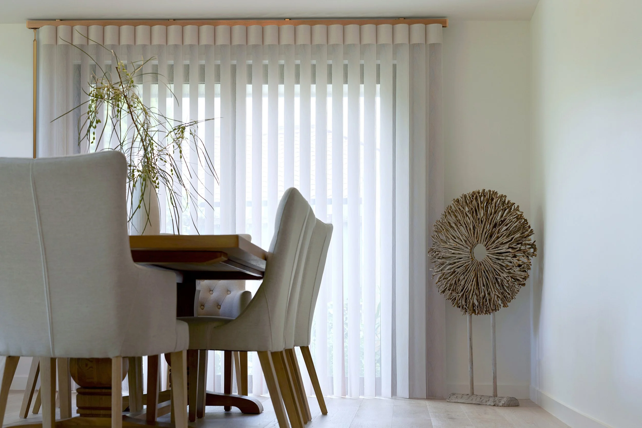 A dining room with a wooden table, beige upholstered chairs, a large plant on the left, sheer vertical blinds on the window, and a decorative wooden sculpture on the right.