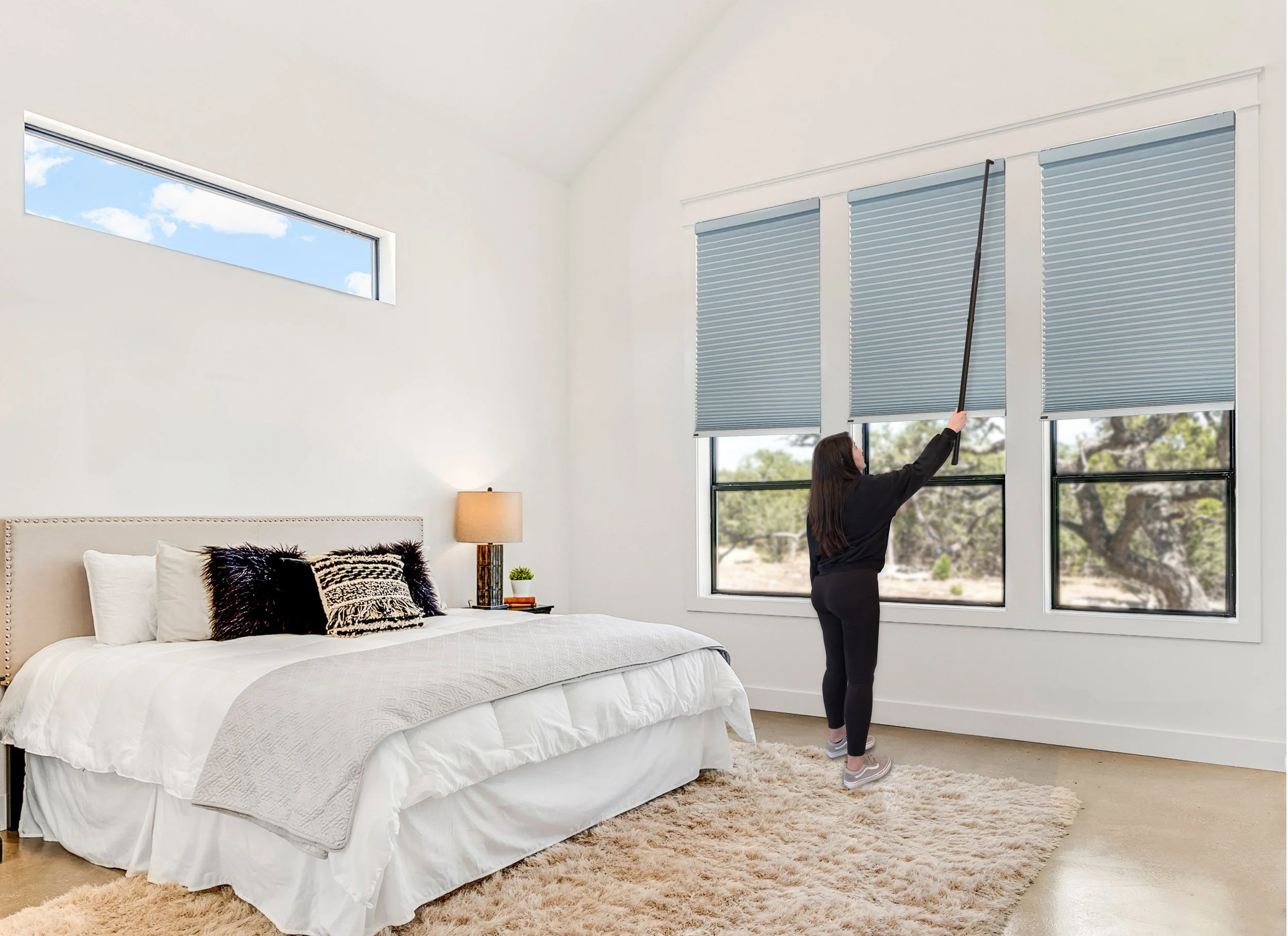 A woman adjusting blue window blinds in a bedroom with a large bed, beige headboard, white bedding, black and animal print pillows, a small bedside table with a lamp, a potted plant, and a beige shaggy rug.