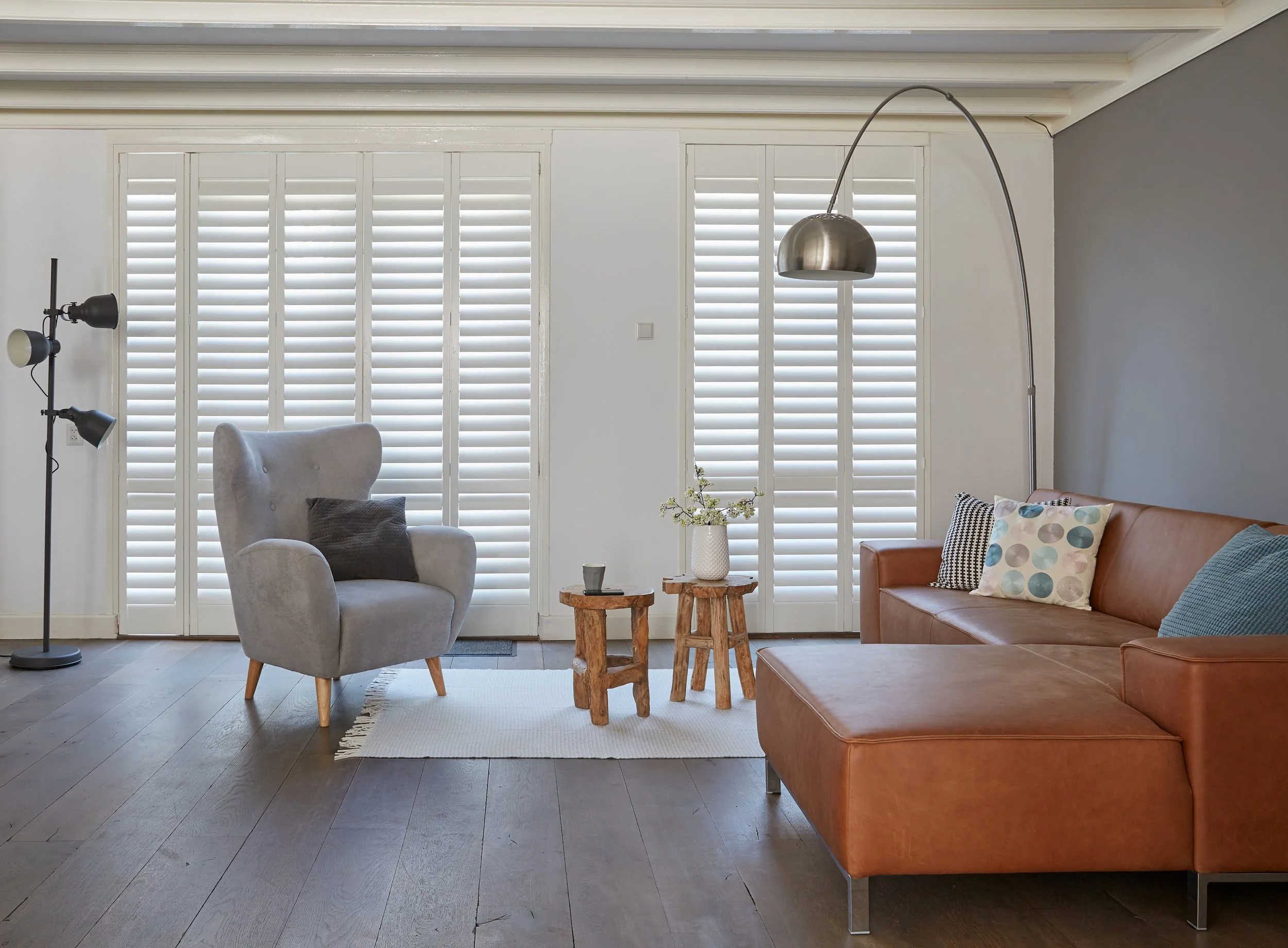 Living room with gray armchair, brown leather sofa, floor lamp, and windows with white shutters