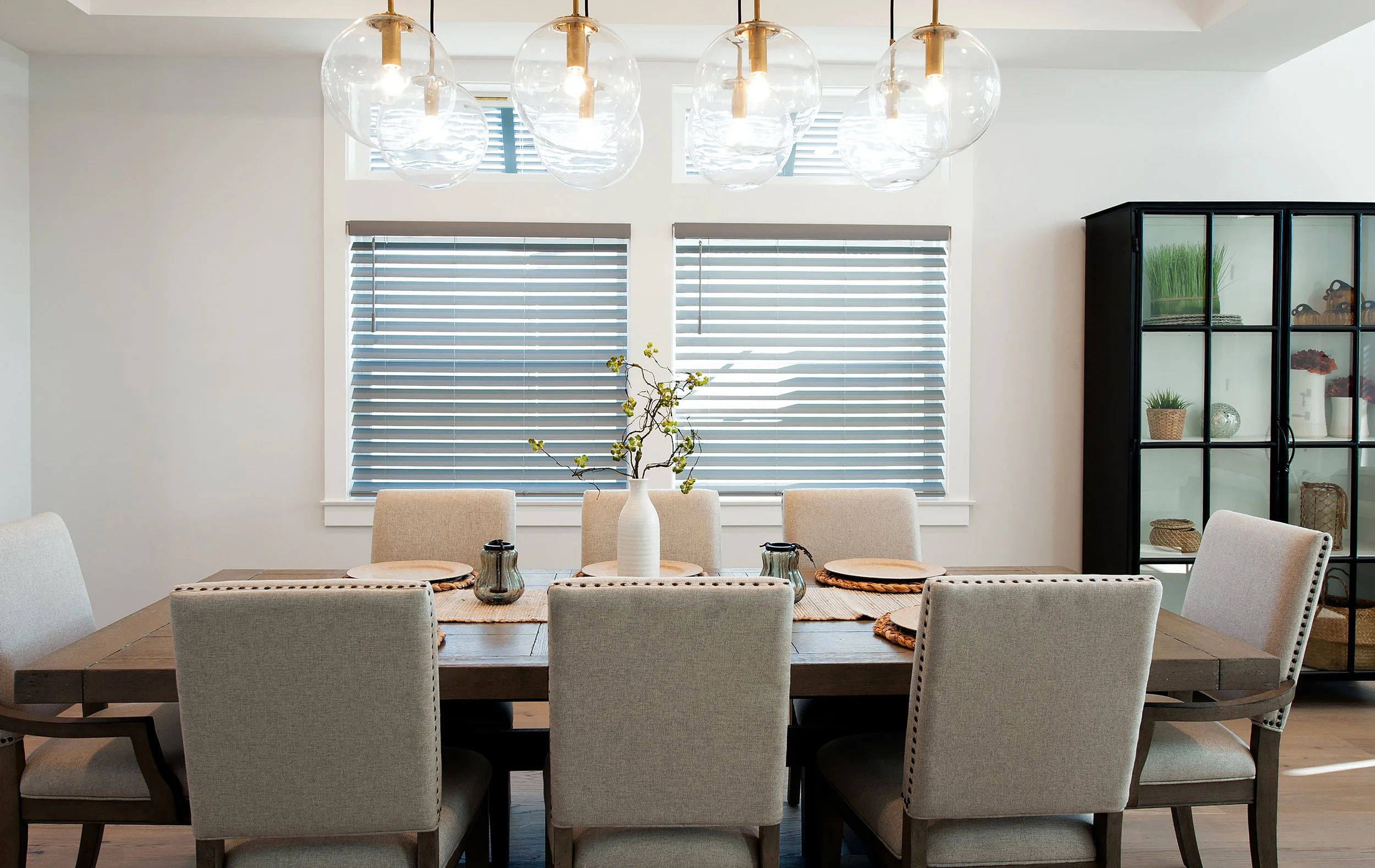 Modern dining room with a wooden table and eight upholstered chairs. A white vase with a branch of yellow flowers is centered on the table. The room has three large windows with horizontal blinds, and a black cabinet with glass doors displaying decor