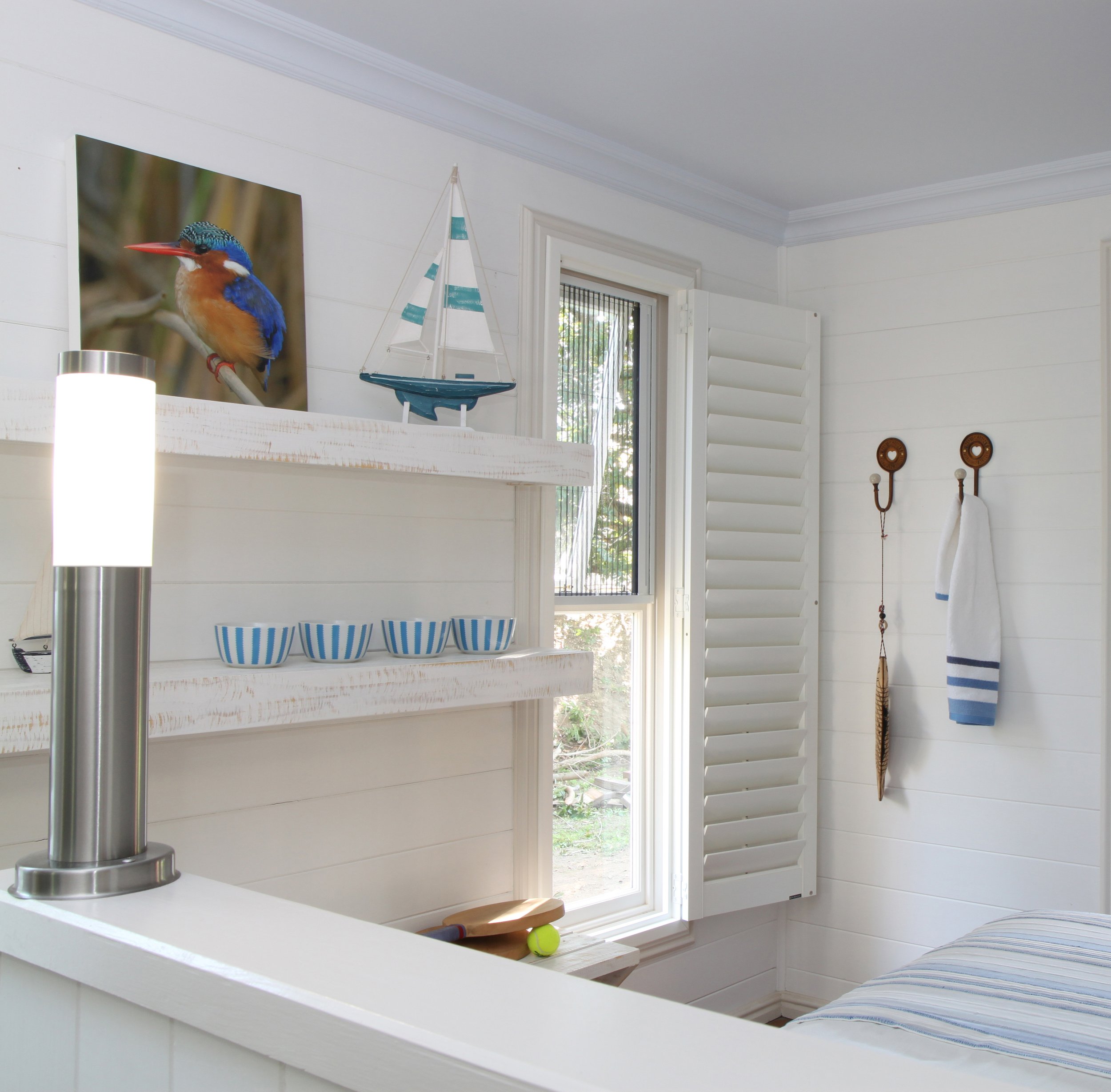 A bedroom corner with white wooden walls and window shutters, a shelf with a colorful bird photograph, model sailboat, and three blue-striped bowls, and wall hooks with a towel and feather decoration.