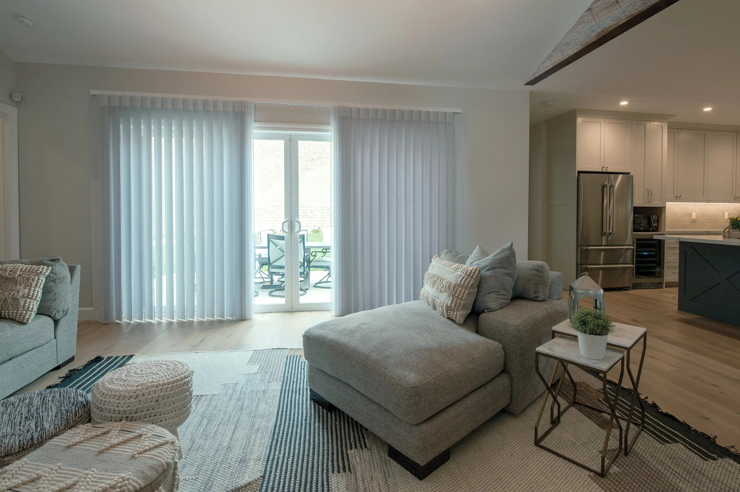 Living room with sliding glass door, beige sectional sofa with pillows, wooden side table with a potted plant and lantern, kitchen with stainless steel appliances in the background, light hardwood floors, and white curtains.