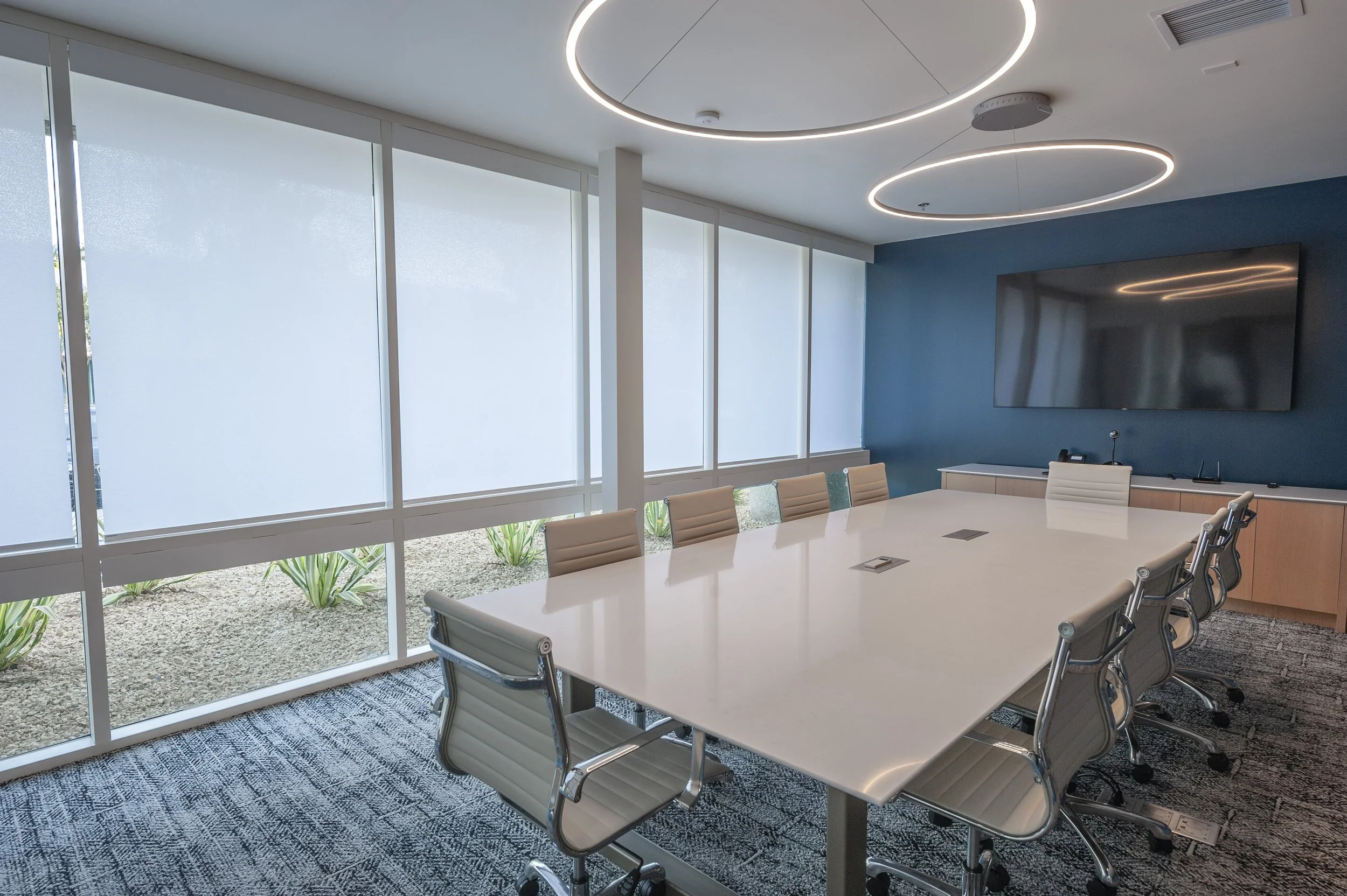 Modern conference room with a large white table, beige chairs, a dark blue accent wall, a mounted TV, large windows with frosted blinds, and circular ceiling lights.