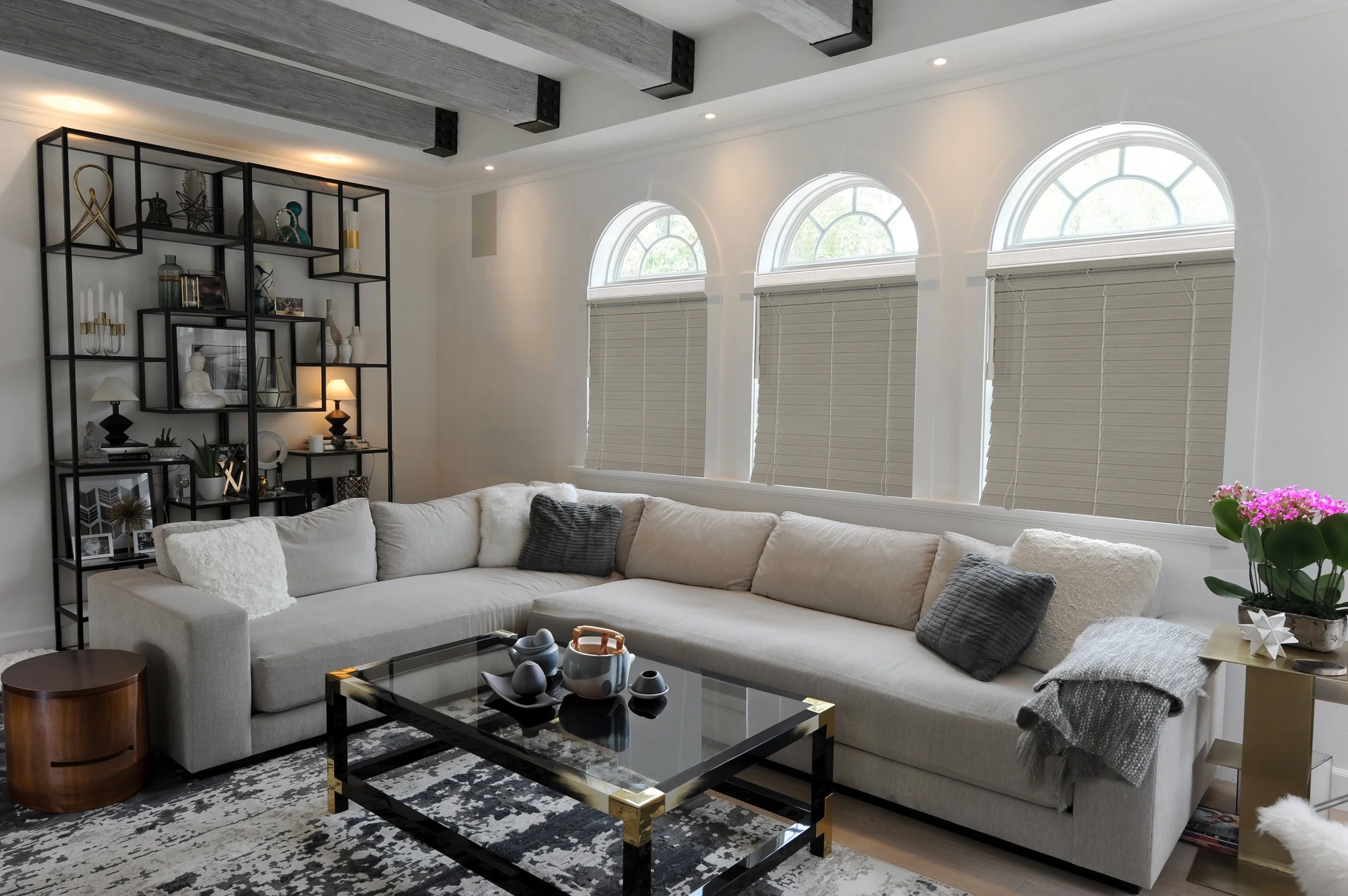 A modern living room with a large beige sectional sofa, a glass coffee table with black and gold accents, a black metal bookshelf with decorative items, potted plants, and arched windows with blinds.