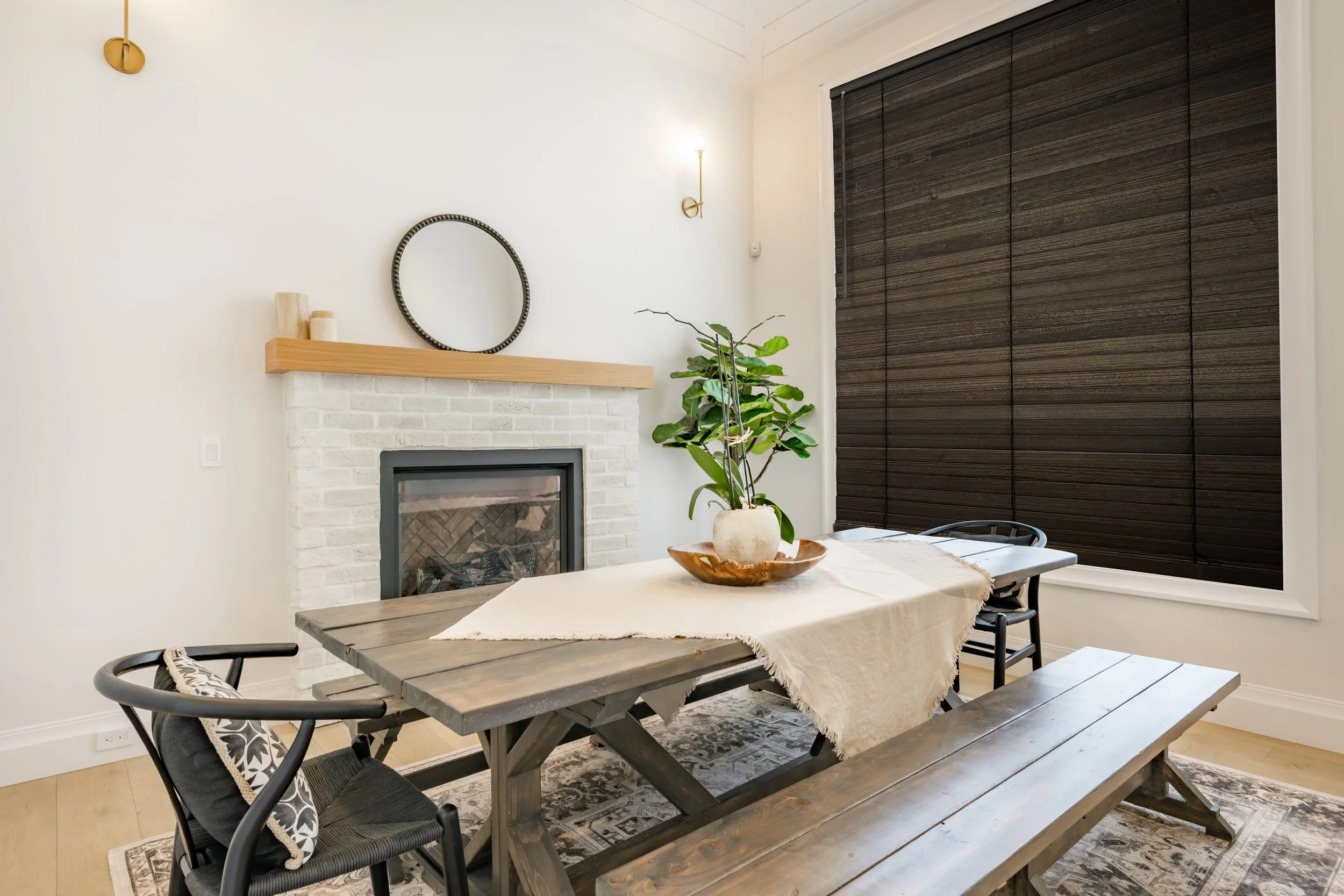 Dining room with wooden table, bench, and black chairs, white brick fireplace with wooden mantel, large potted plant, dark window blinds, and decorative mirror on white wall.