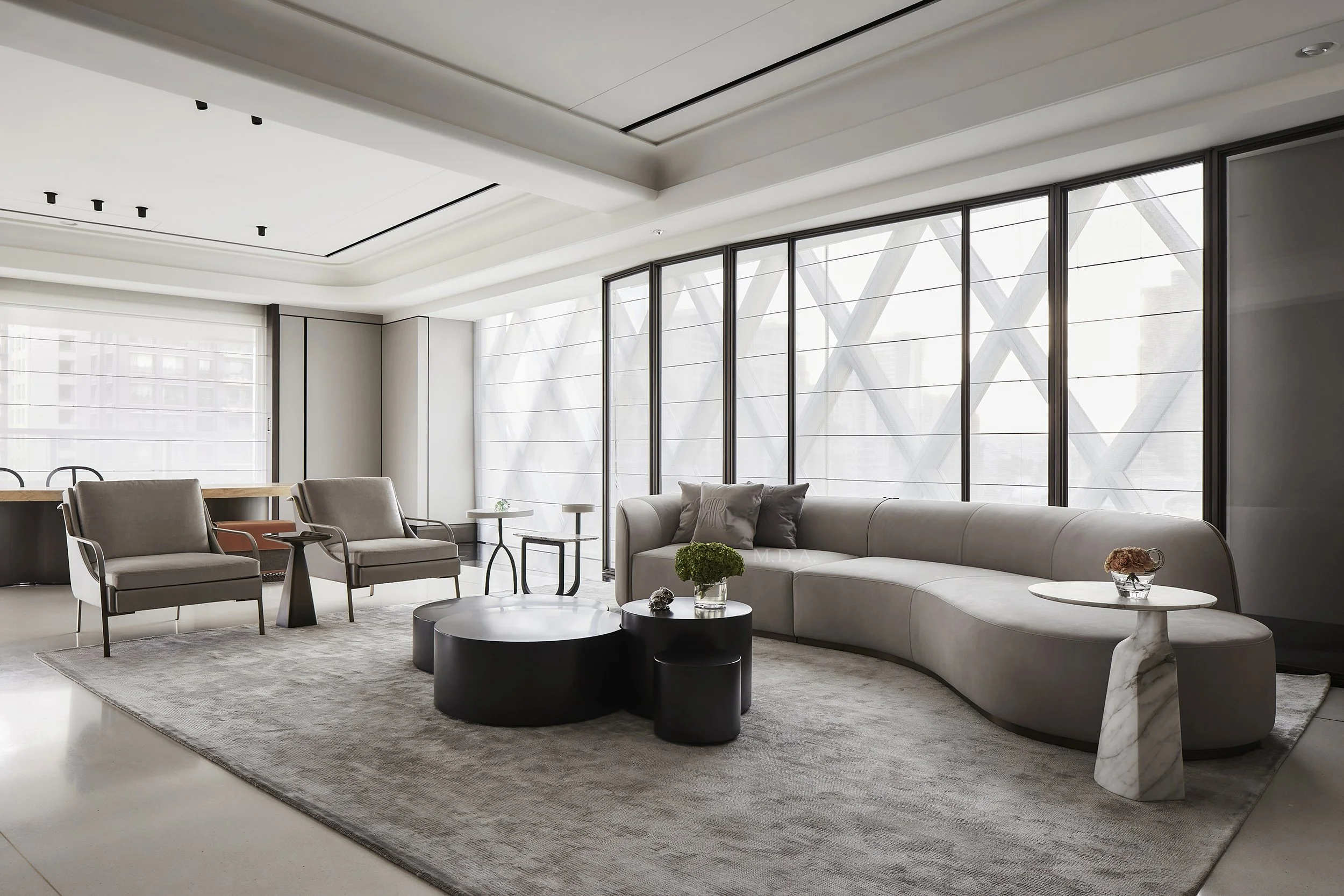 Modern living room with gray curved sofa, two gray armchairs, black coffee tables, marble side table with flowers, and large windows with city views.
