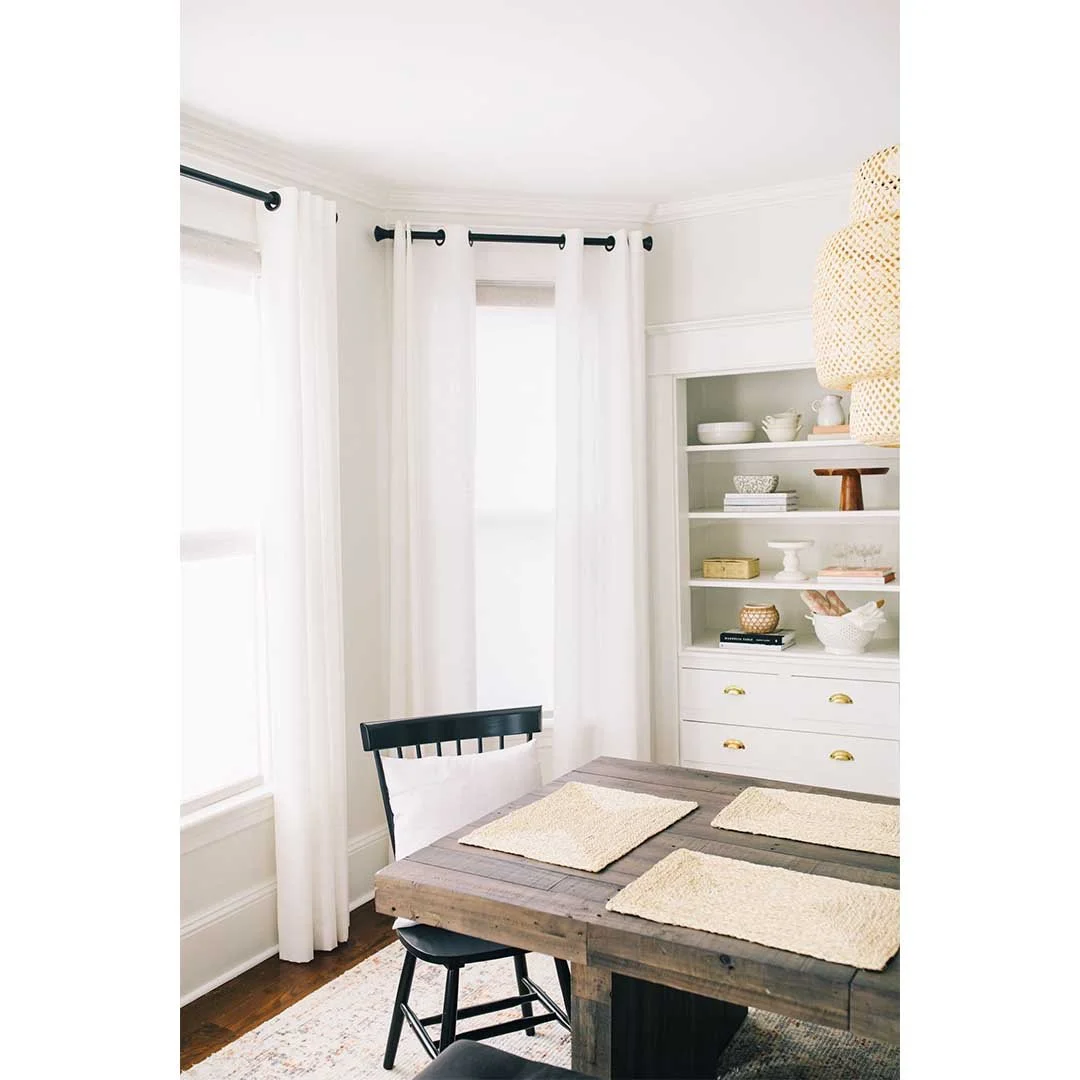 A dining room with white curtains, a wooden table with three beige placemats, a black chair, and a white built-in shelf with decorative items and bowls.