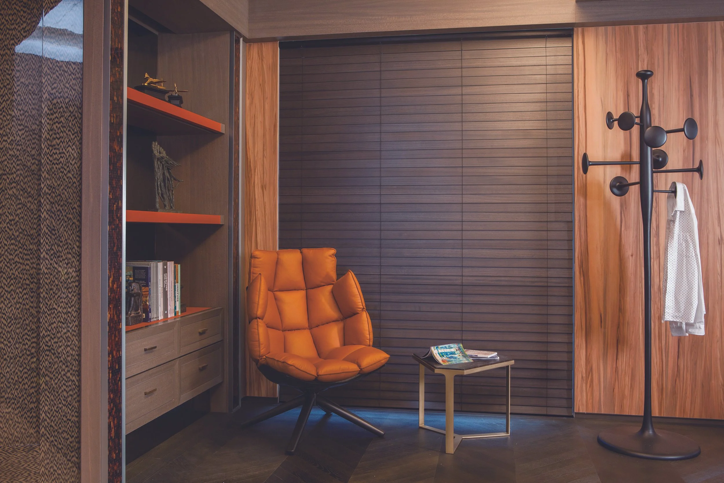 A cozy reading corner with a padded orange armchair, a small wooden side table with magazines, a black coat rack with a white shirt, and built-in wooden shelves filled with books and decorative items, set against wooden wall paneling and blinds.