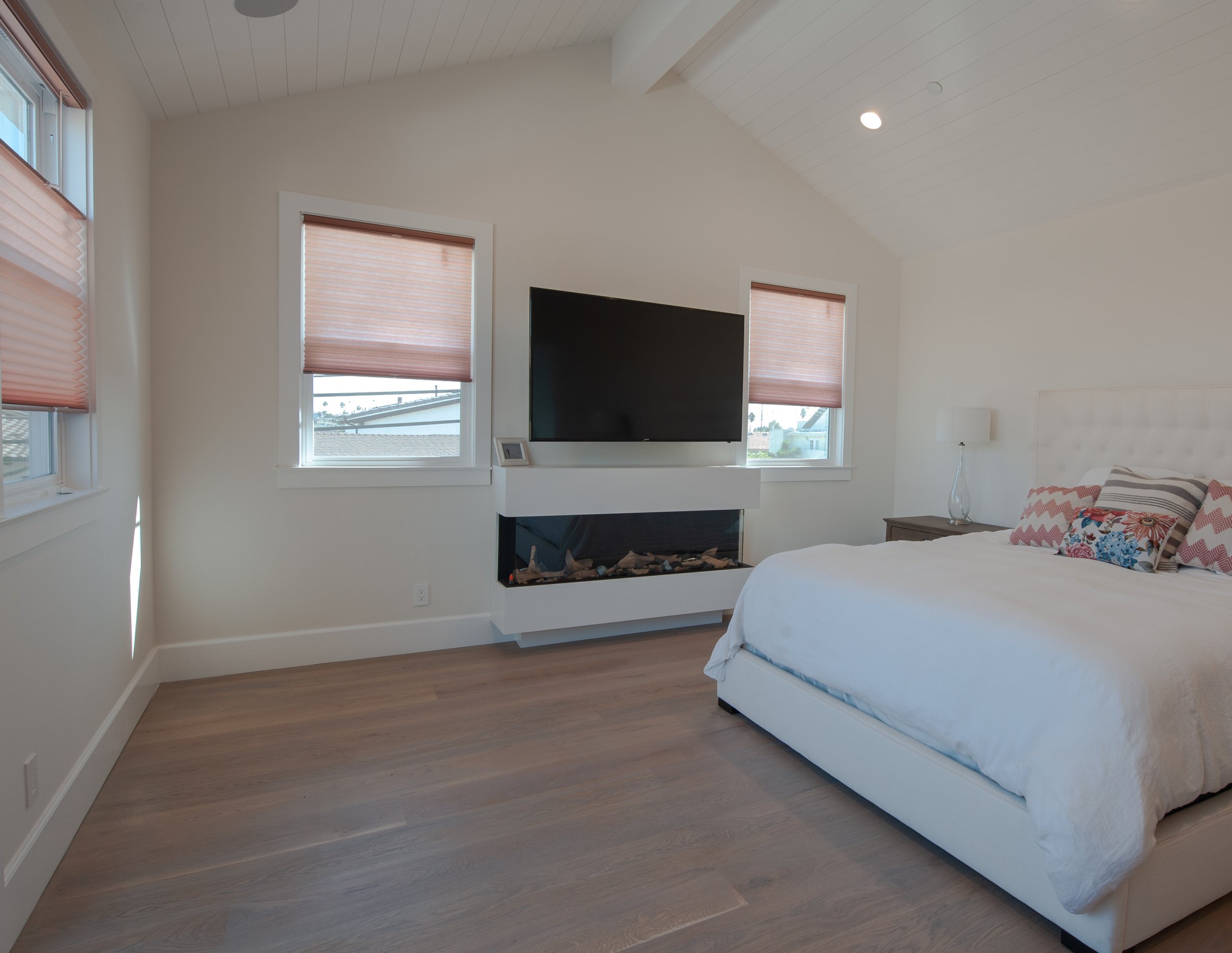 A bedroom with a white bed, decorative pillows, a bedside table with a lamp, a TV mounted above a modern fireplace, and three windows with pink blinds.
