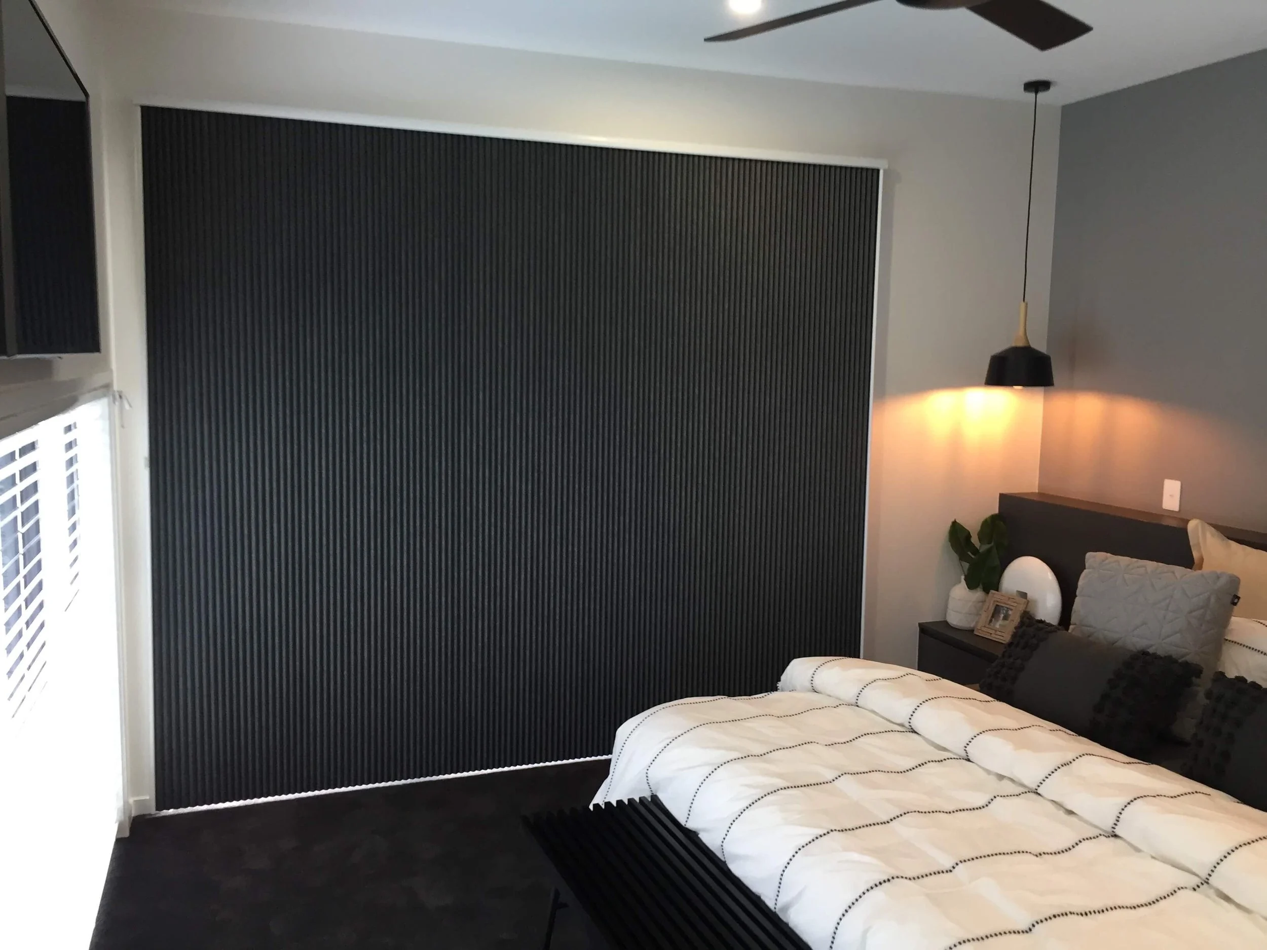 Modern bedroom with black textured accent wall, white bedding with black lines, bedside table with decorative items, and a black pendant light.
