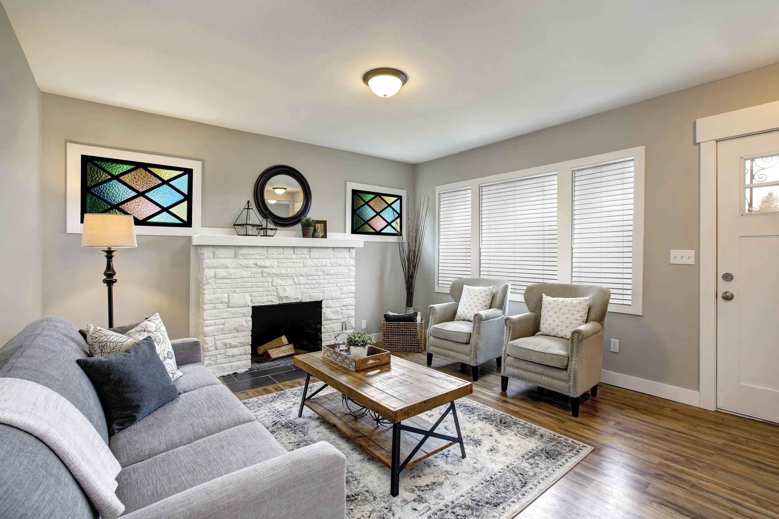Living room with a gray sofa, two beige armchairs, a wooden coffee table, and a white brick fireplace, modern decor, hardwood flooring, and windows with white blinds.
