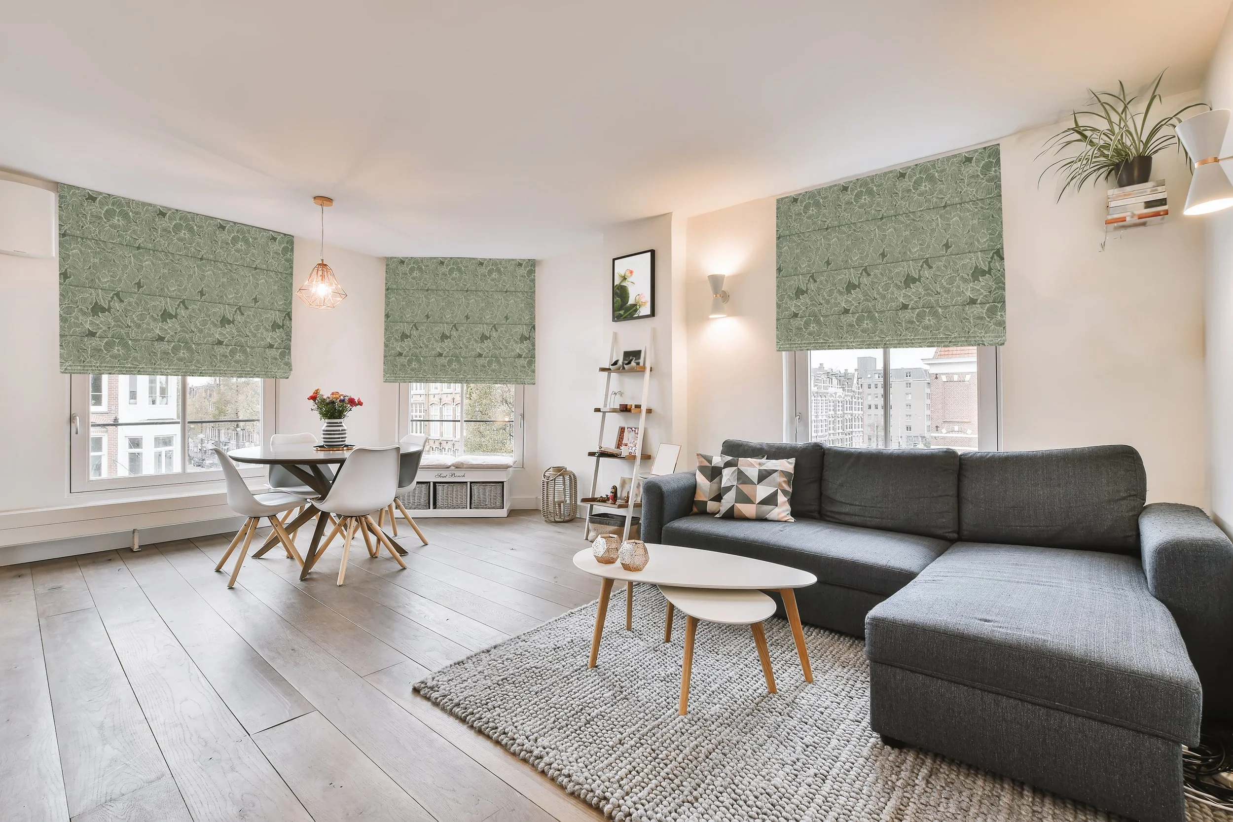 Bright living room with large windows, green window shades, California-style light wood flooring, a gray sectional sofa with patterned pillows, a white coffee table, a tall white bookshelf, and decorative plants.