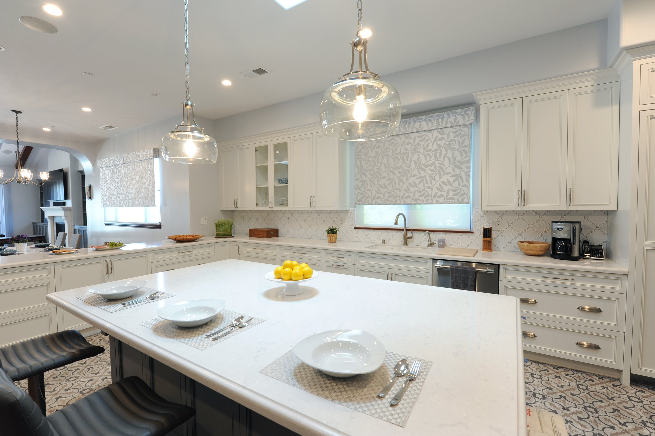 Modern kitchen with white cabinets, a large island, pendant lights, window shades, and utensils on the countertop.