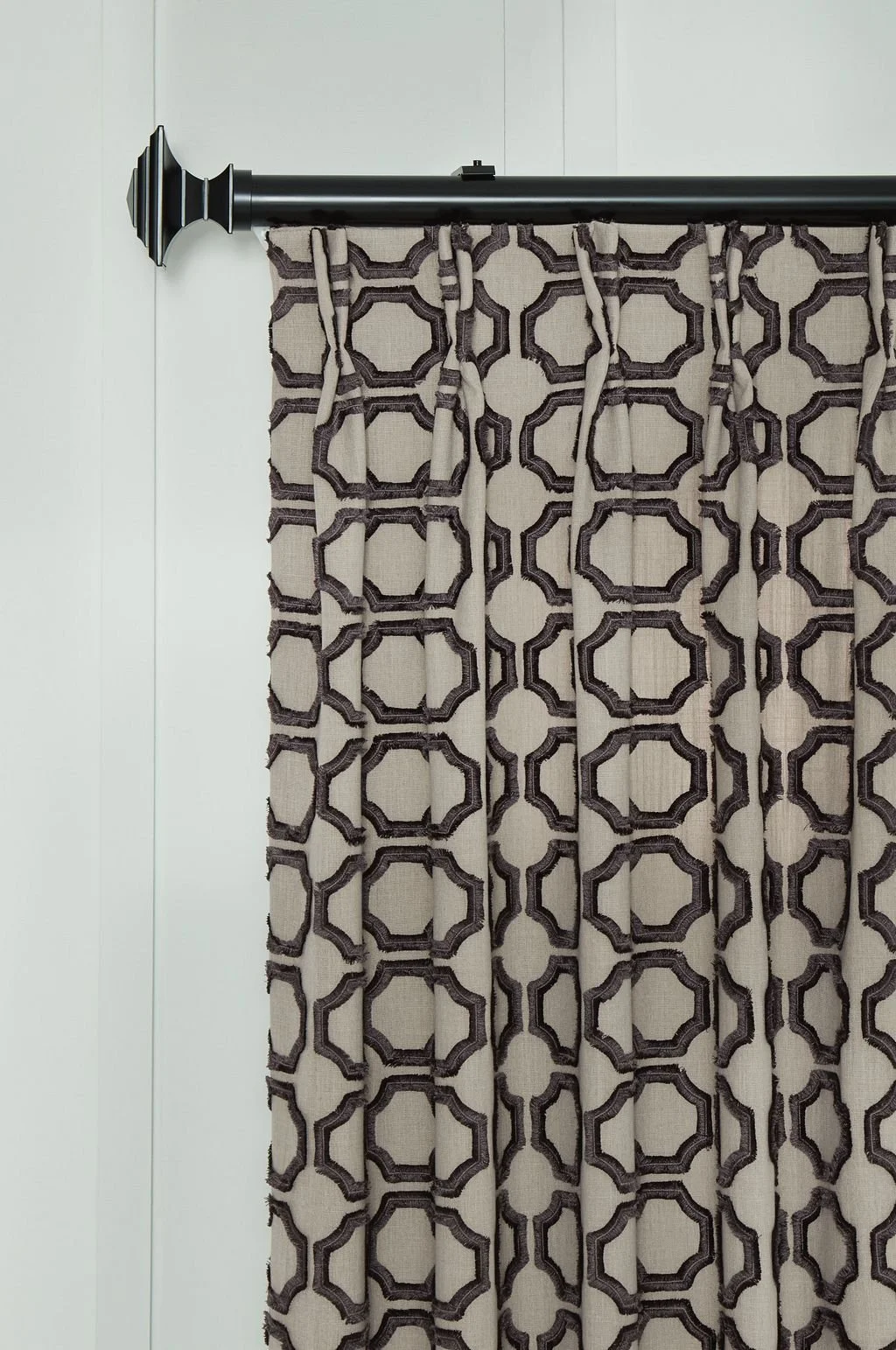 A beige curtain with a dark brown geometric pattern hanging from a black curtain rod with decorative end brackets, mounted to a white wall.