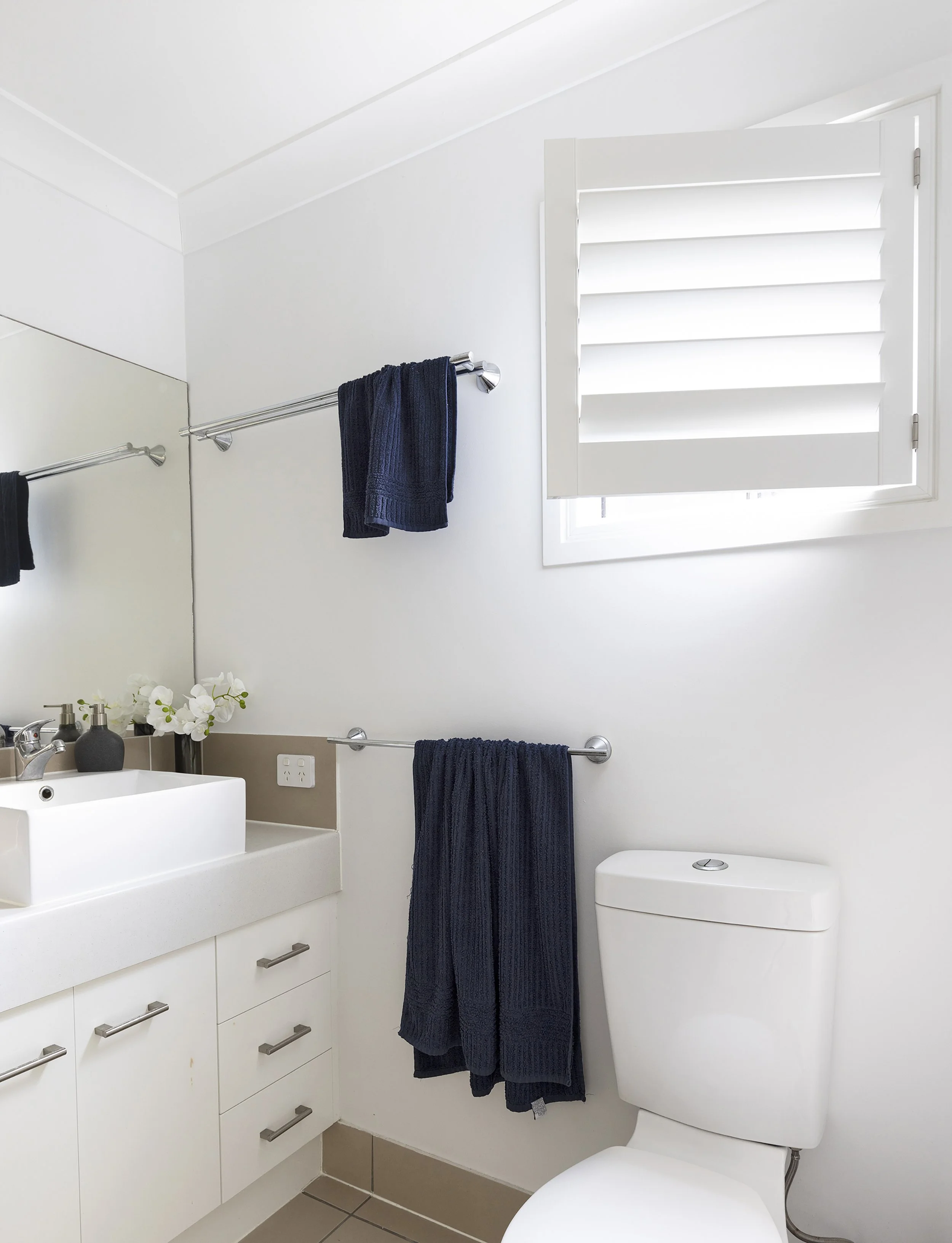 A minimalist bathroom with white walls, a white vanity with silver handles, a square vessel sink, and a large mirror. There are navy blue towels on a silver towel rack, a small black soap dispenser, and a white vase with flowers. The bathroom has a b