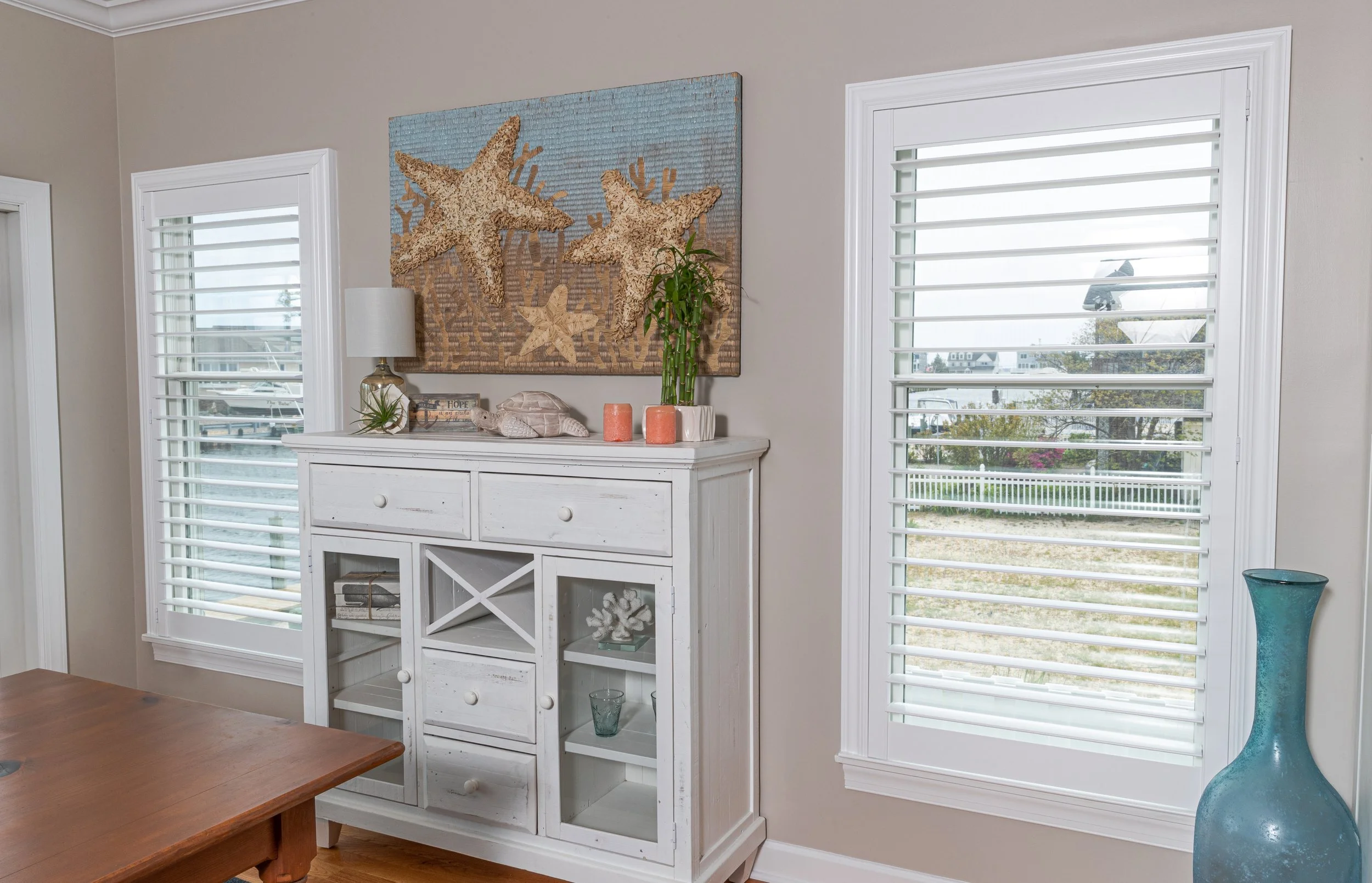 Beach-themed decor on a white wooden cabinet with starfish artwork above, two windows with white shutters, a table partially visible in the foreground, and a tall blue glass vase on the floor to the right.