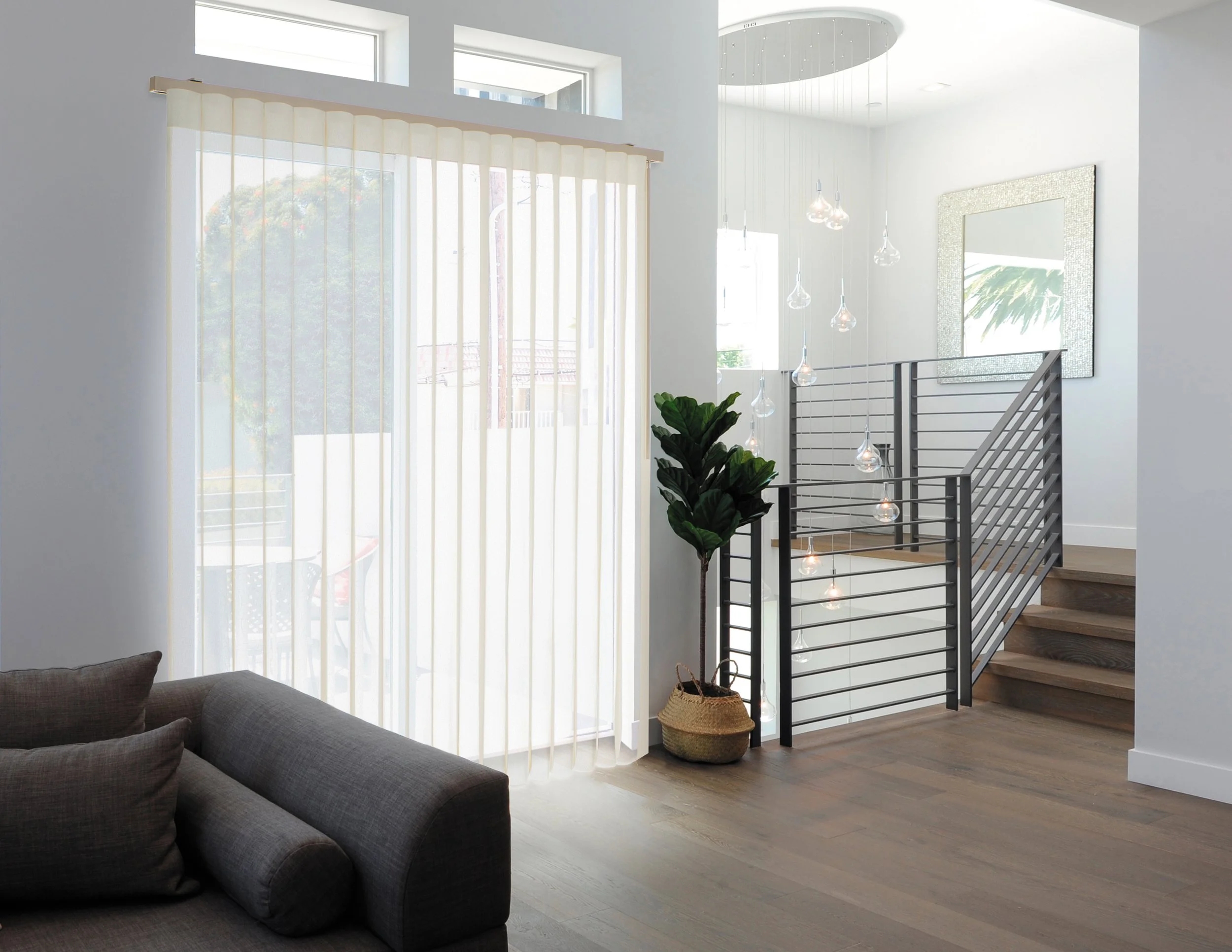 Interior view of a modern living space with a staircase, a large window with vertical blinds, a potted plant, framed artwork, and a glimpse of outdoor scenery.
