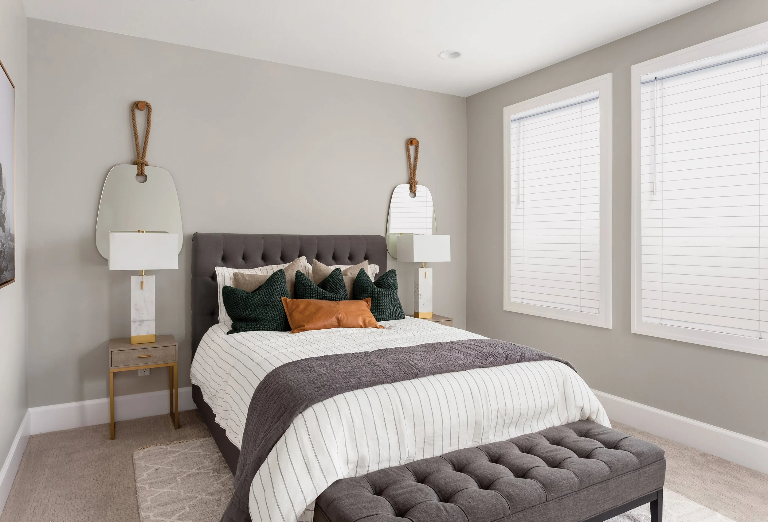 A bedroom with a gray upholstered bed, black and beige pillows, and a white and gray striped blanket. Two white nightstands with lamps and mirrors with hanging ropes are on each side of the bed. The room has two large windows with white blinds, gray 