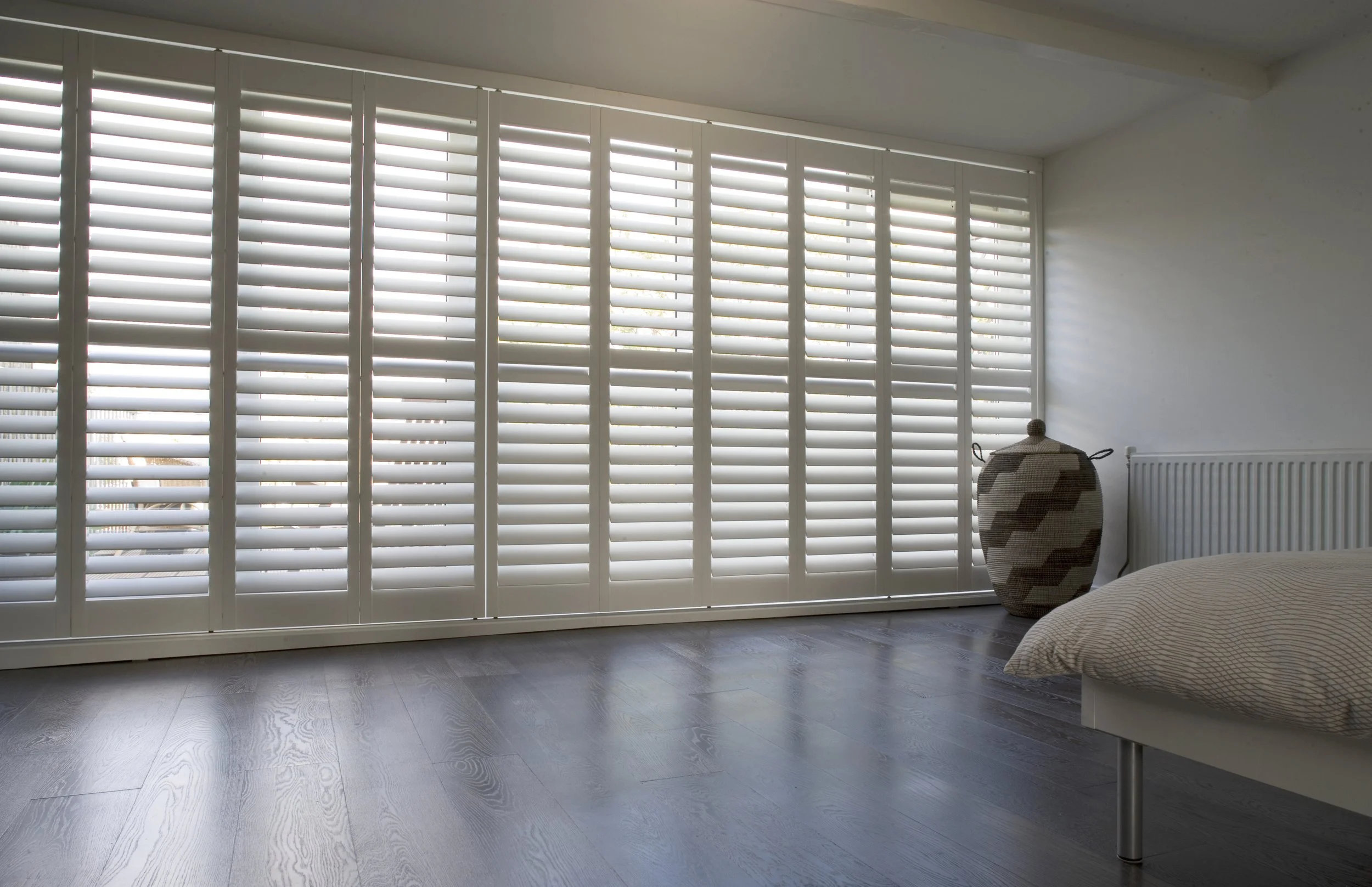 A bright bedroom with large white plantation shutters covering a window or sliding door, a wicker vase near the wall, and part of a bed with a beige pillow and light-colored bedding.