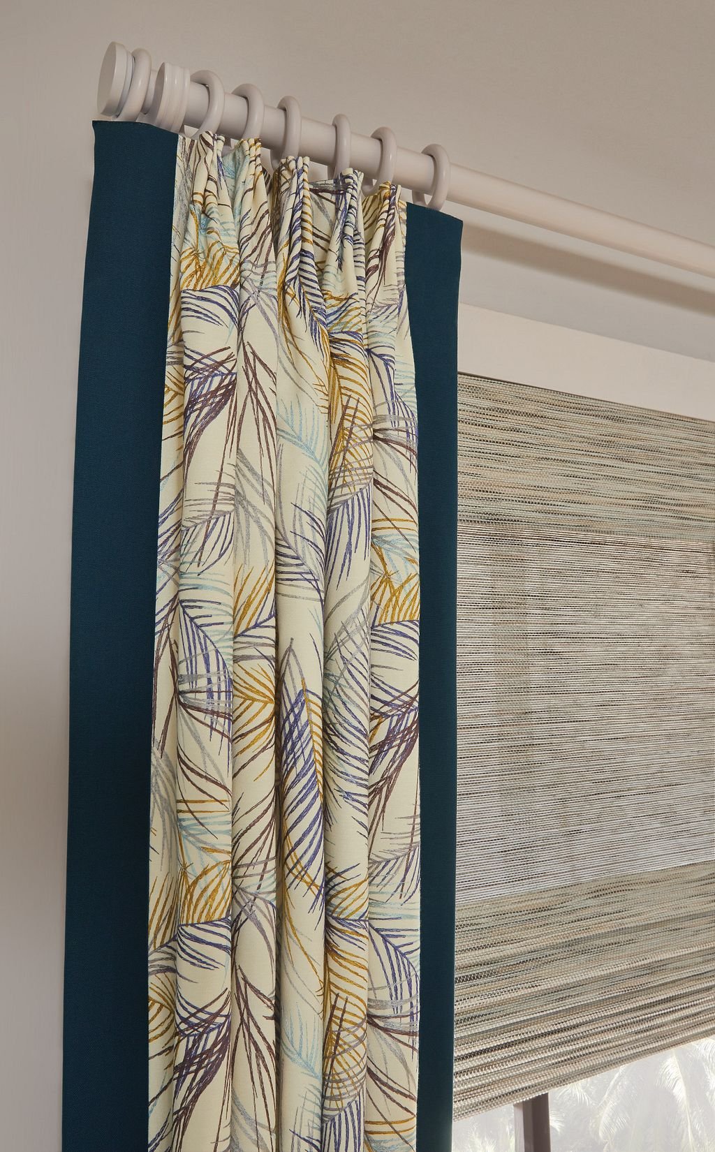 Close-up of curtains hanging from a white curtain rod, with a patterned fabric with leaf design and a Roman shade with horizontal stripes, in a room with light-colored walls.