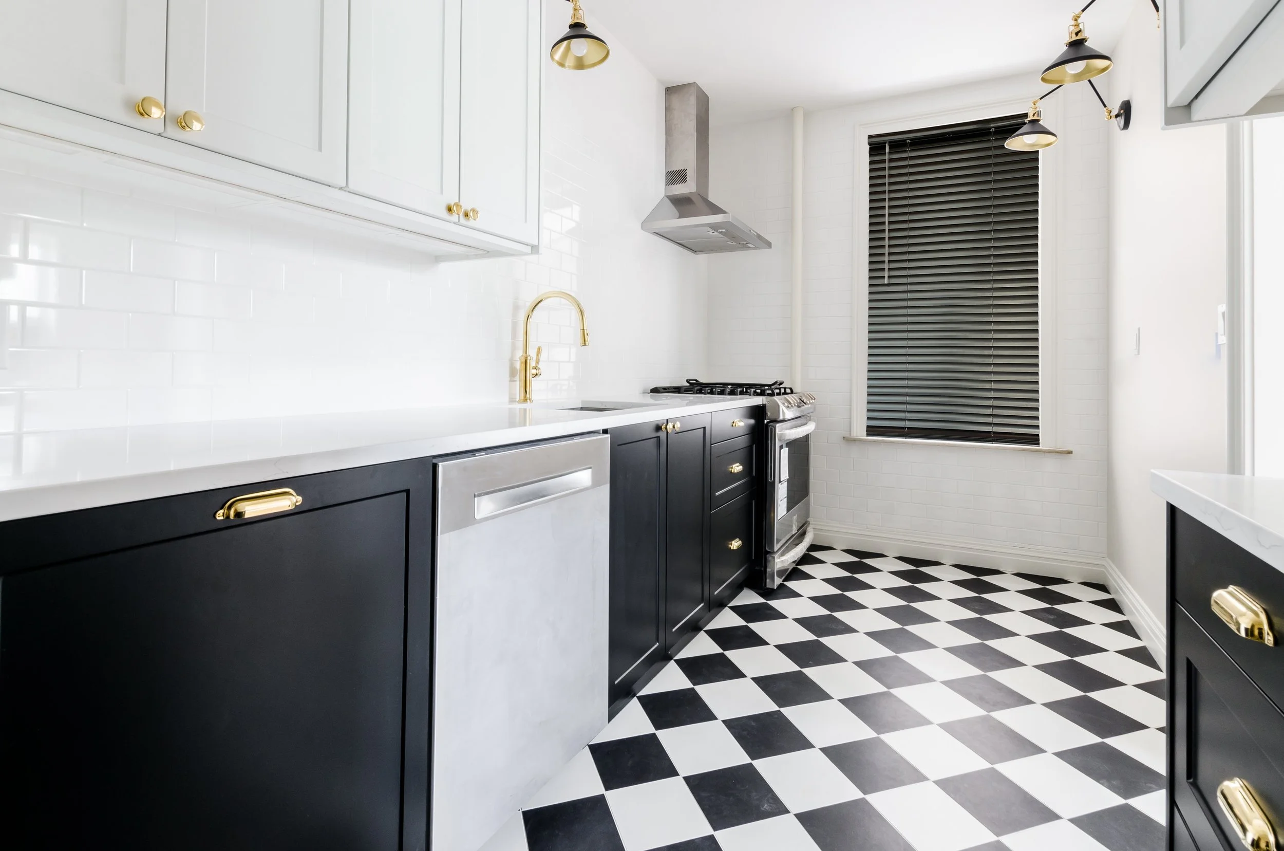 Modern kitchen with black and white cabinetry, gold hardware, checkered black and white floor, white subway tile backsplash, stainless steel dishwasher, stove, and range hood, black window blinds.