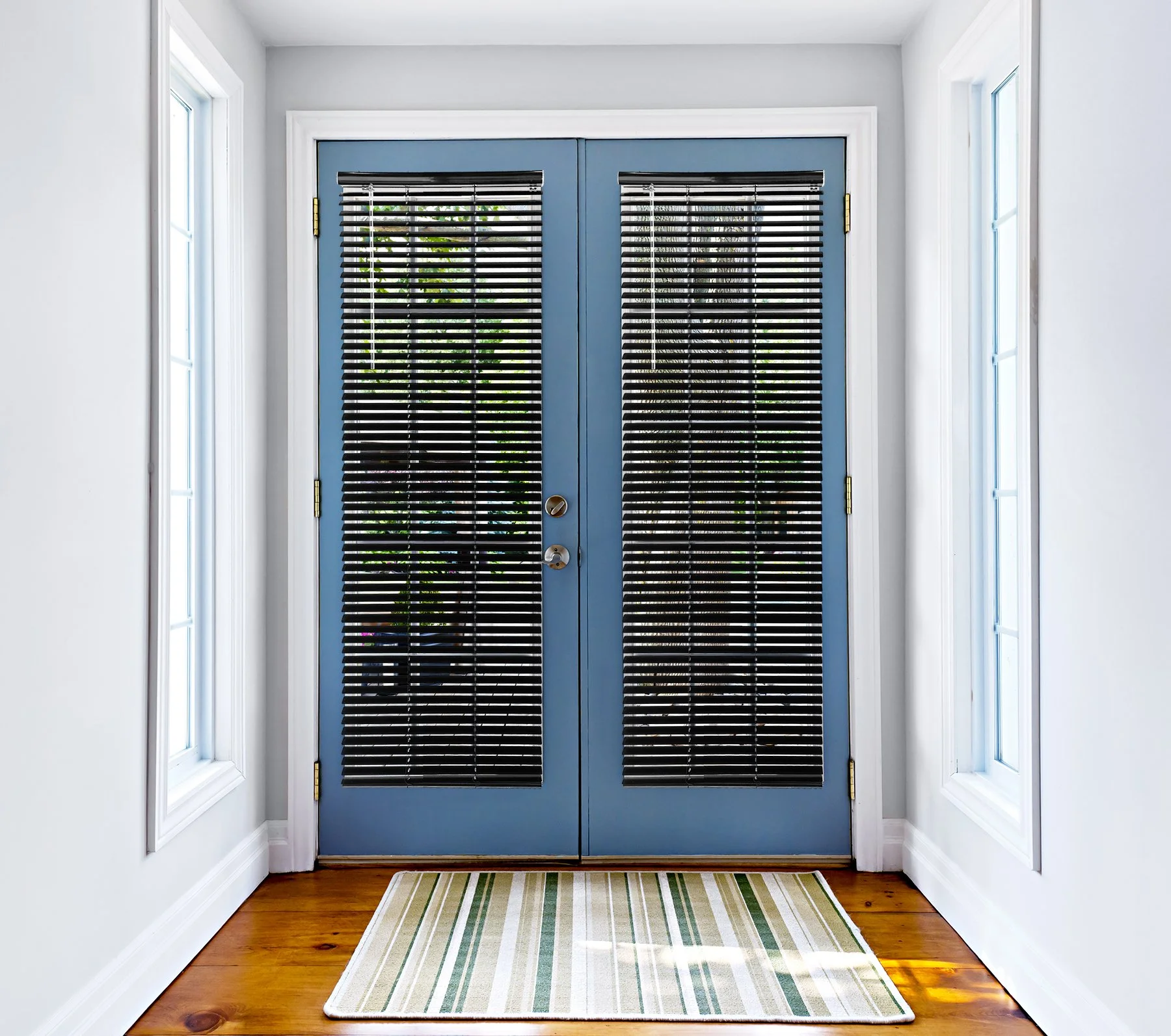 Blue double doors with black blinds, white window frames on both sides, hardwood floor, striped rug in front.