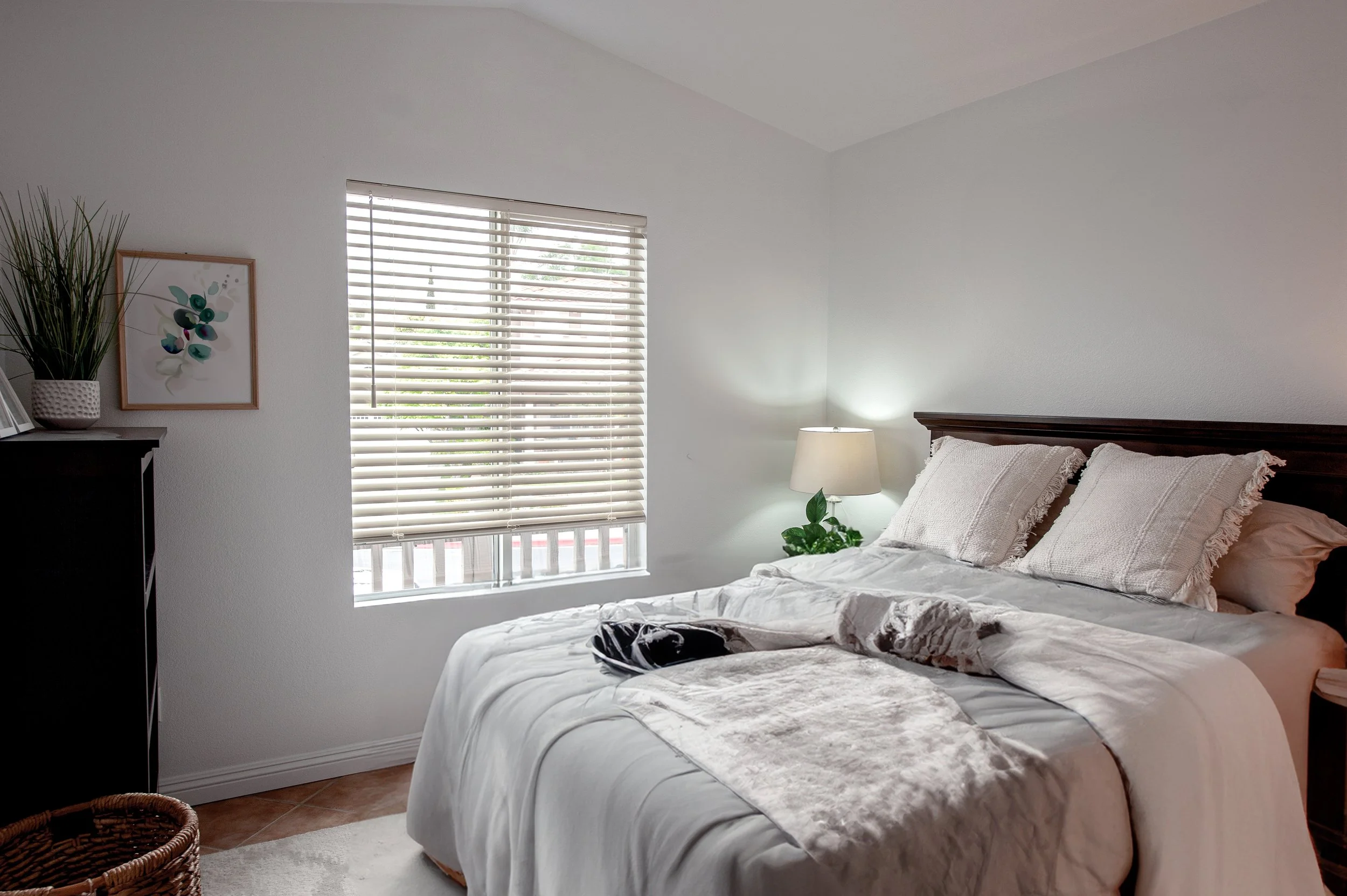 A neatly made bed with white bedding and pillows in a bright bedroom. There's a window with blinds, a bedside lamp, and some indoor plants and decorative items.