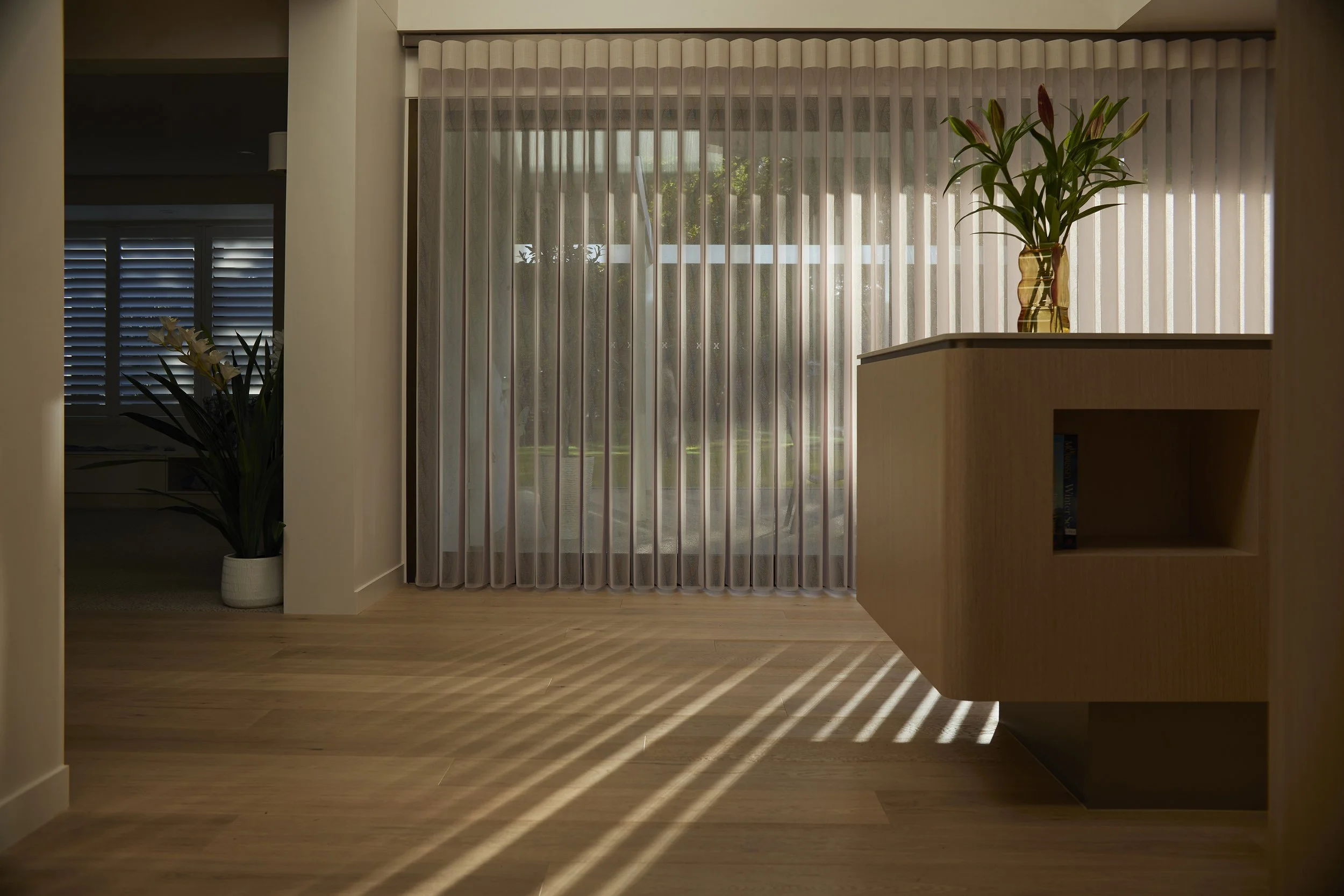 Living room with wood flooring, sheer curtains covering large windows with sunlight streaming through, and potted plants on a wooden cabinet and in the corner.