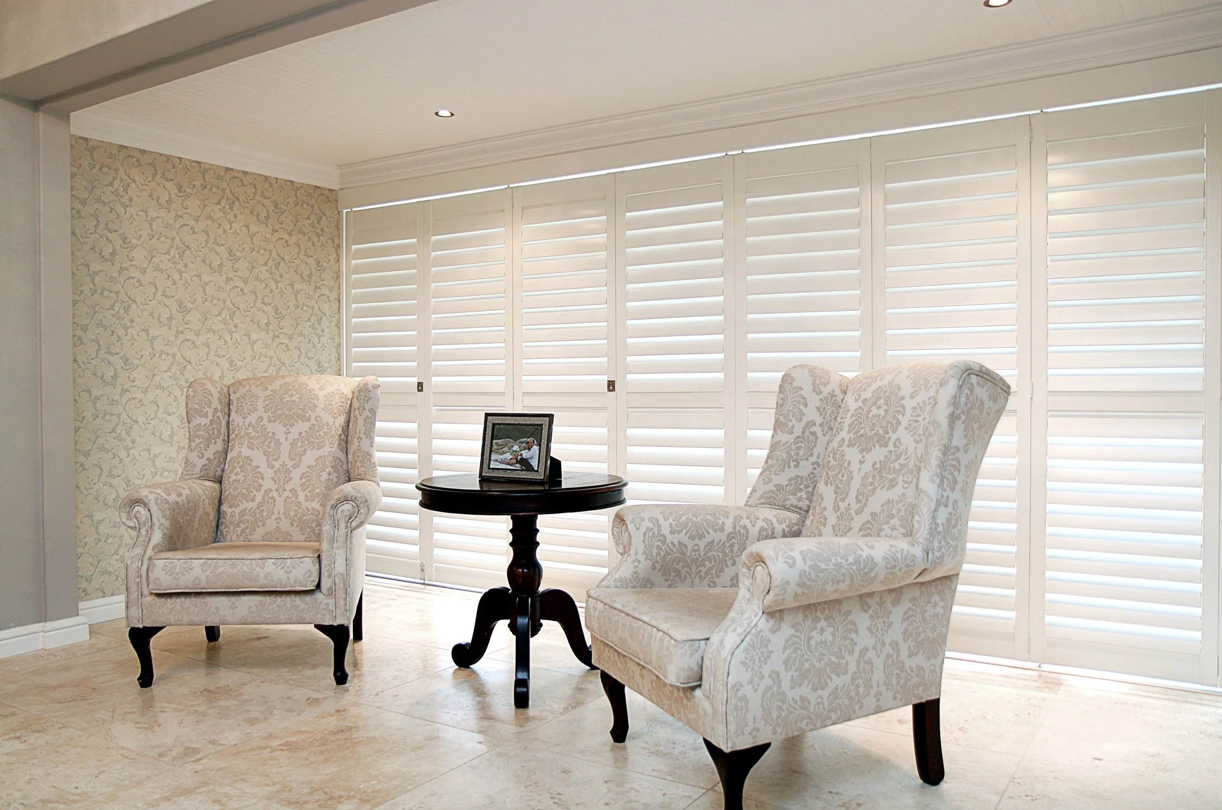 Two armchairs with floral upholstery facing each other, separated by a small round table with a photo frame on it. Large windows with white plantation shutters in the background and beige tiled flooring.