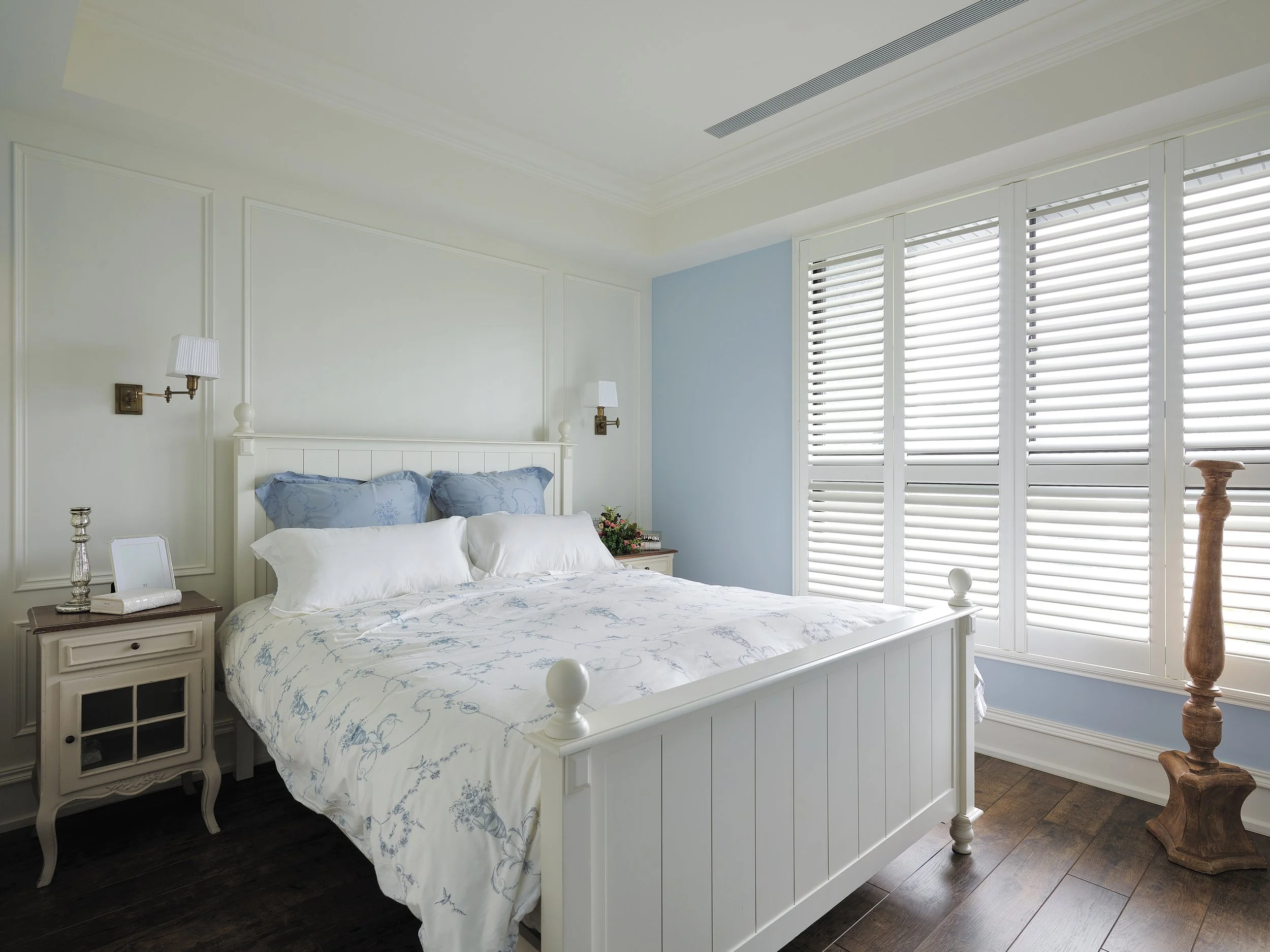 Bright bedroom with a white bed, blue pillows, white bedding, two nightstands, and a large window with white shutters.
