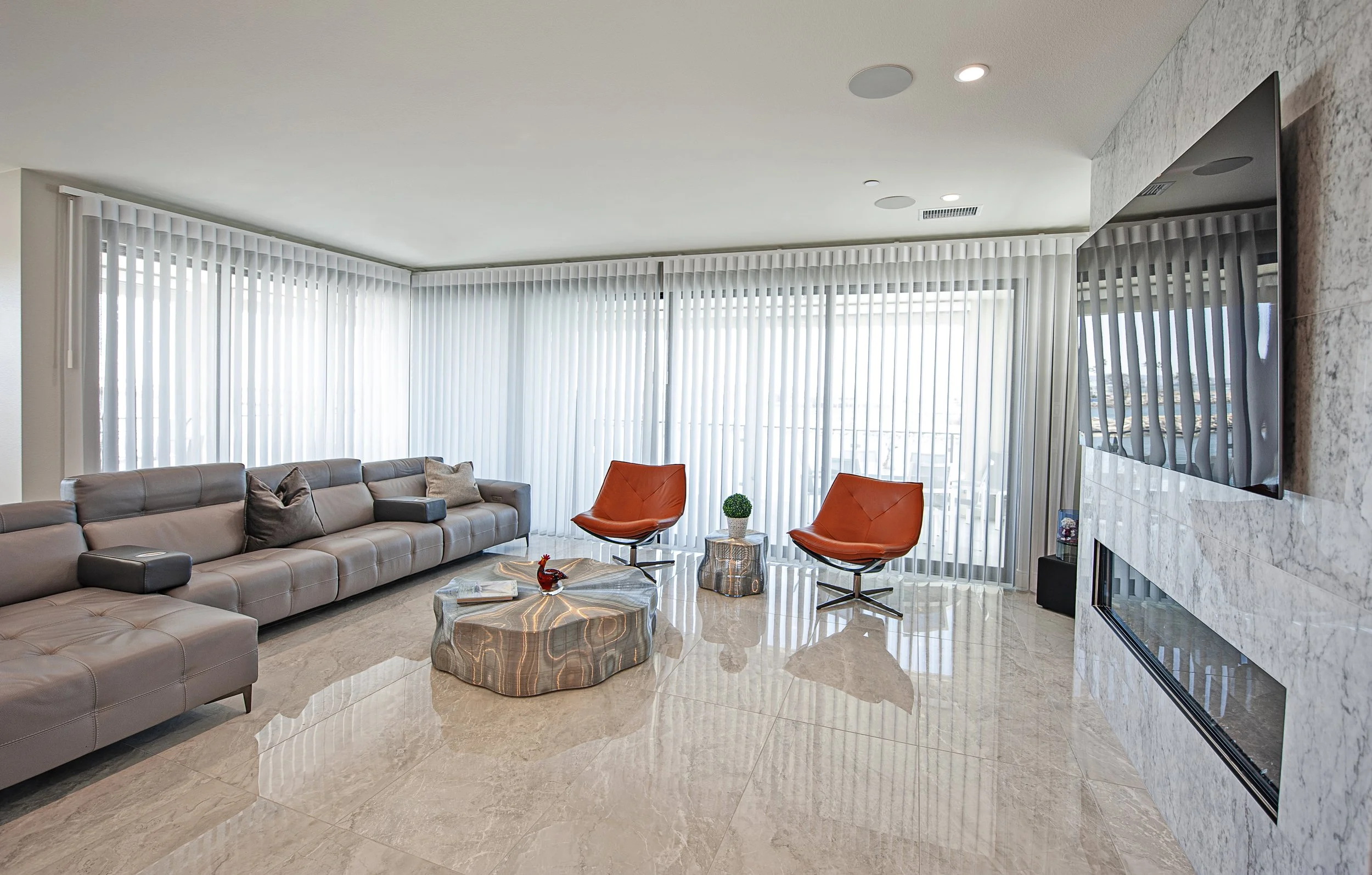 Modern living room with beige tiled floor, a large light gray sectional sofa with cushions, two orange chairs, a small side table with a small plant, a unique wooden coffee table, and a wall-mounted TV with a fireplace below, tall windows with vertic
