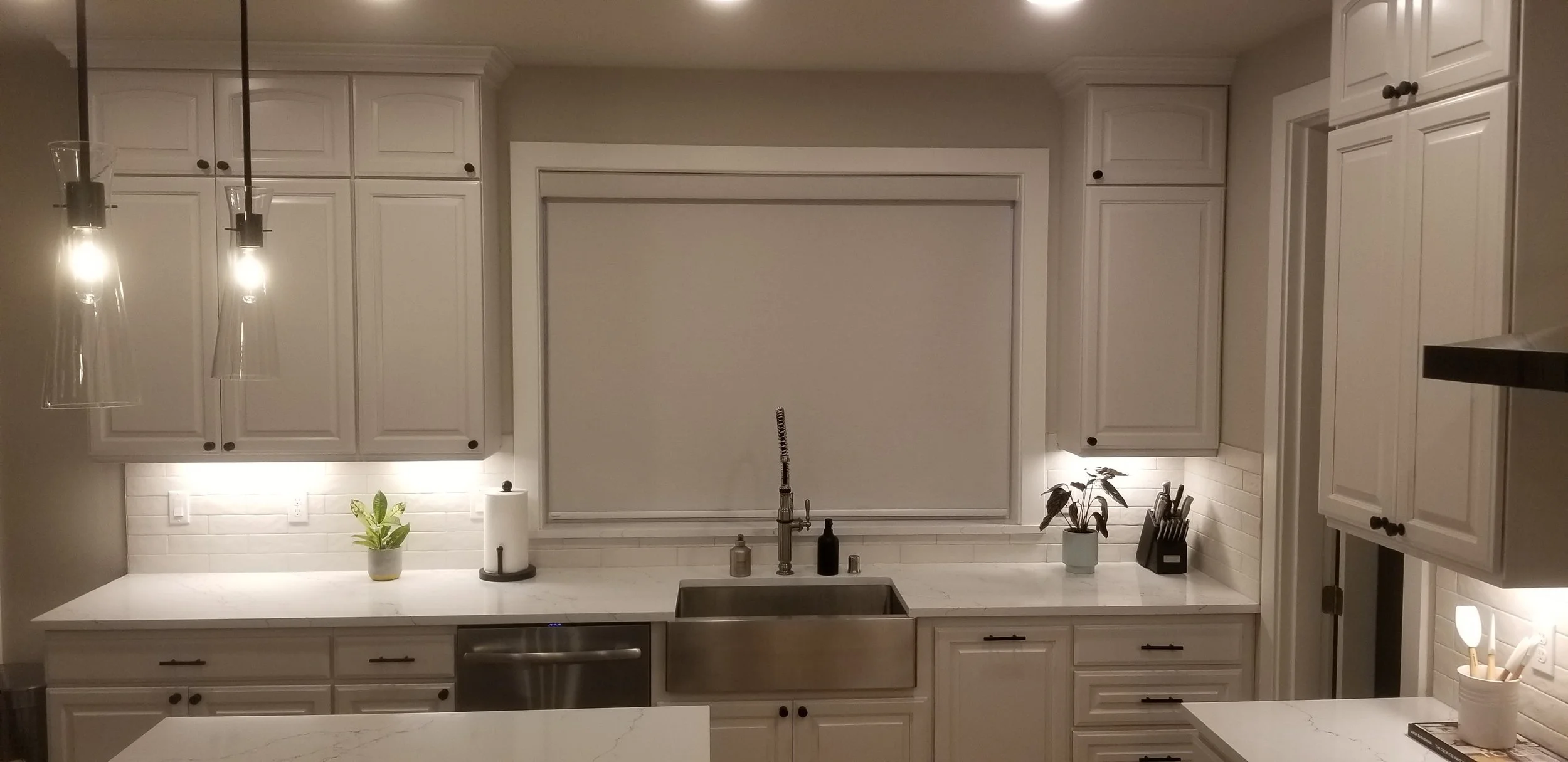 Modern kitchen with white cabinets, white marble countertops, stainless steel farmhouse sink, black faucet, potted plants, paper towel holder, knife block, and pendant lights.