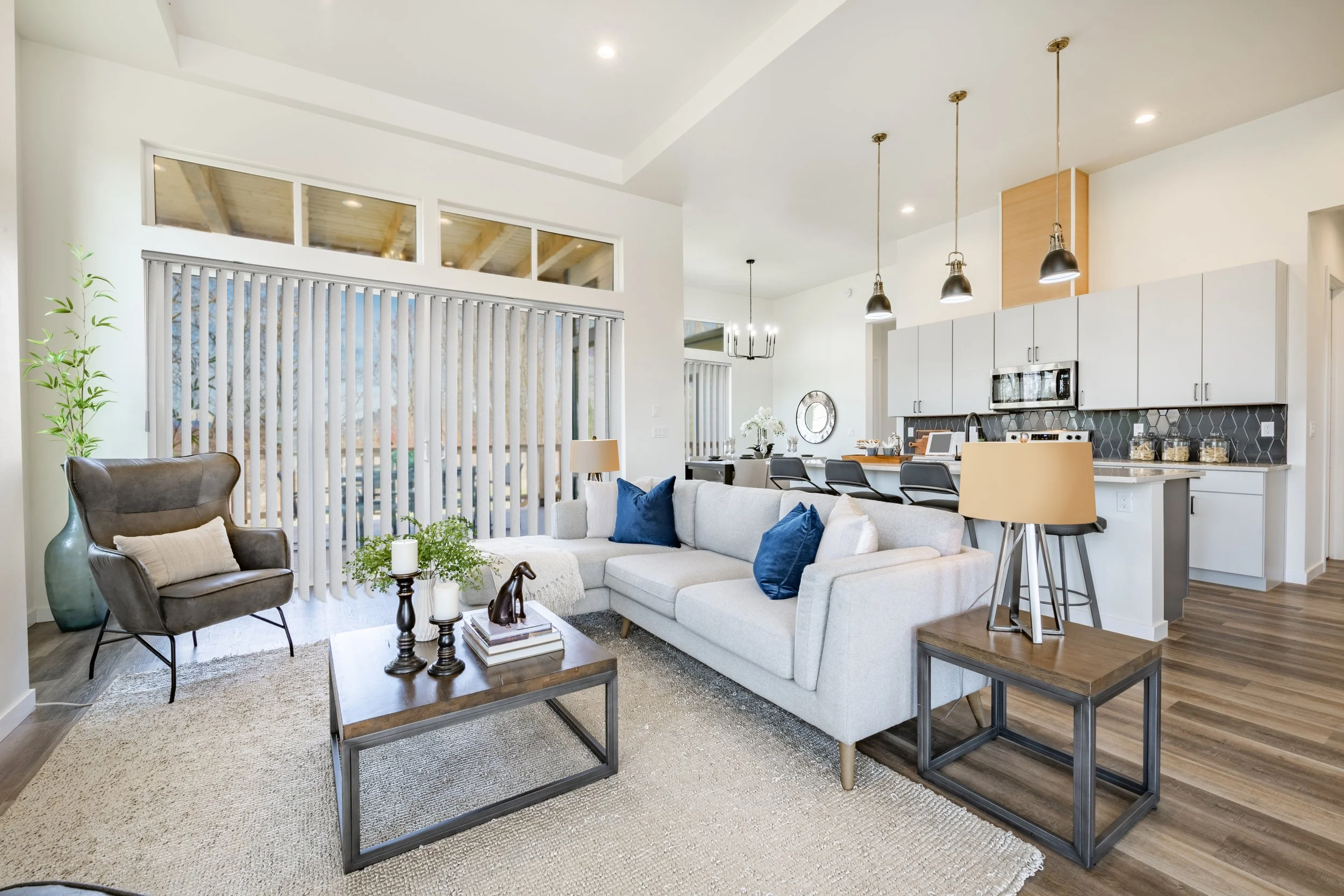 Bright, modern living room with white sofa, blue pillows, dark armchair, glass coffee table, large vase, and sliding glass door with vertical blinds separating from the kitchen and dining area.