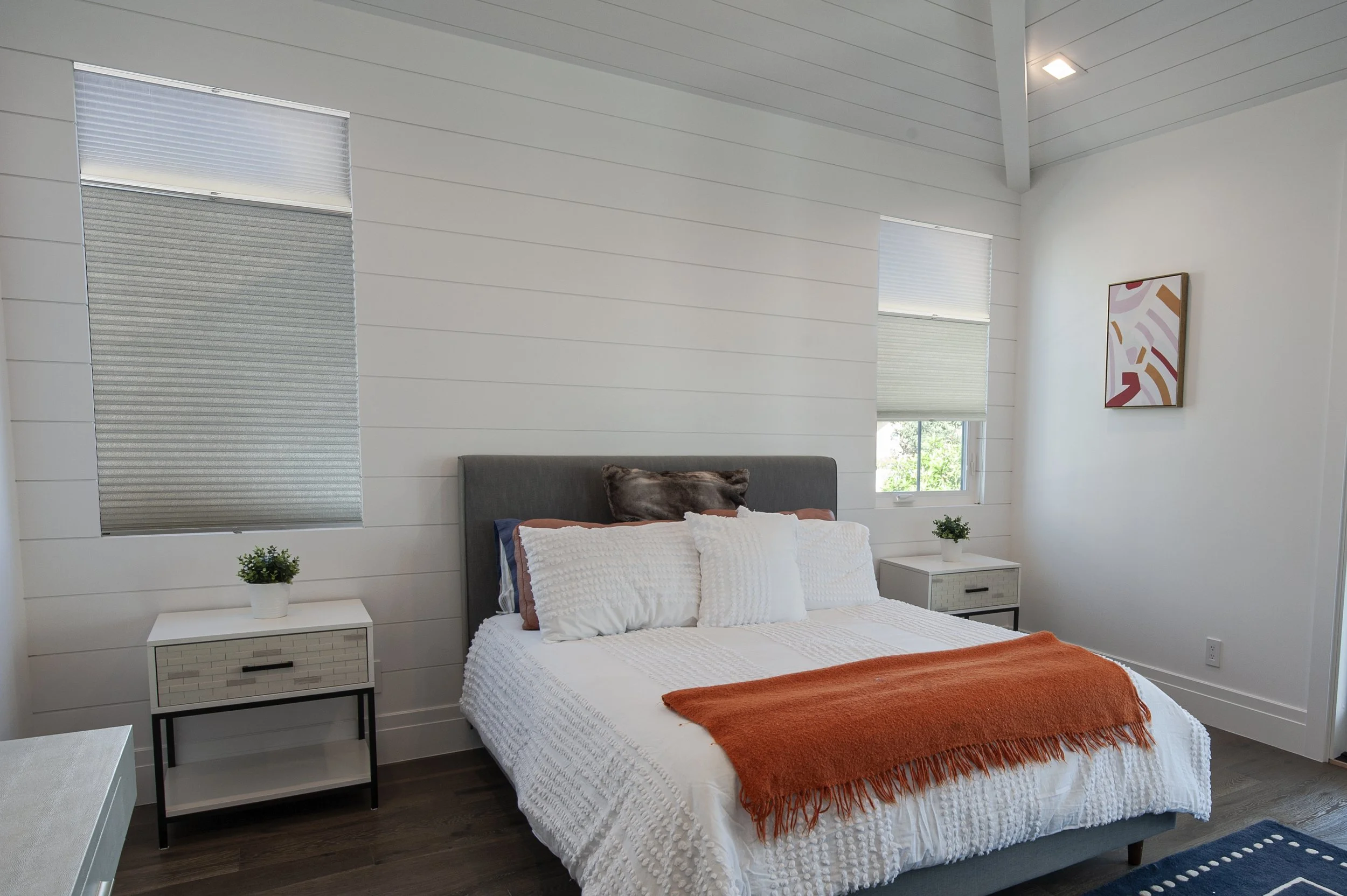 A modern bedroom with a bed that has white bedding, an orange throw blanket, and multiple pillows. There are two white nightstands with potted plants on each side. The wall behind the bed is painted white with horizontal paneling, and there are two w