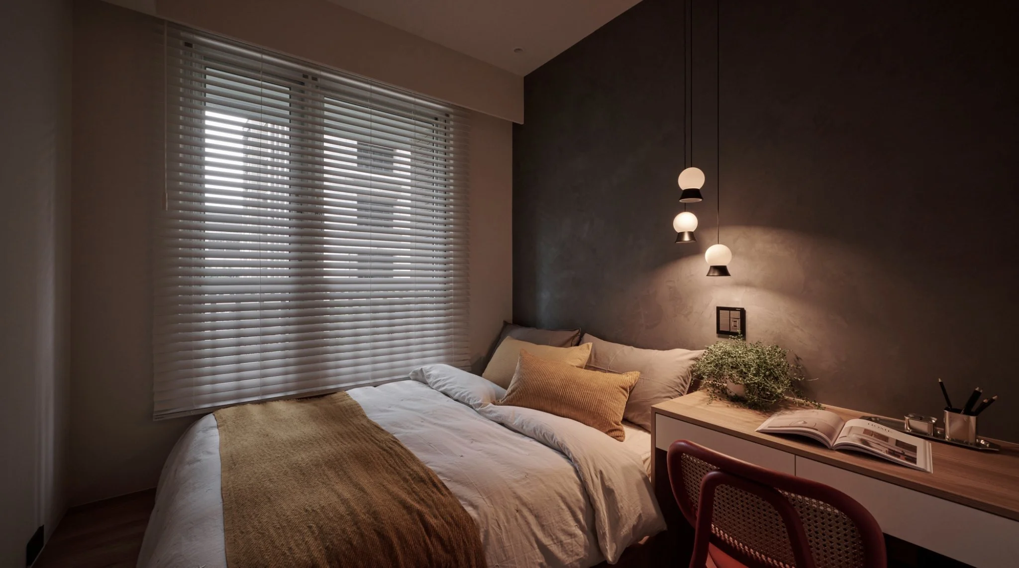 A modern bedroom with a large window, blinds, a bed with beige and gray pillows, a tan blanket, and a wooden desk with an open book, a plant, and some stationery.