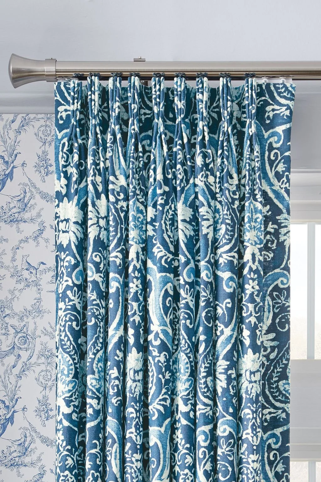Blue patterned curtains hanging on a silver curtain rod in front of a window with white blinds, with wallpaper featuring a blue floral and animal motif on the wall to the left.