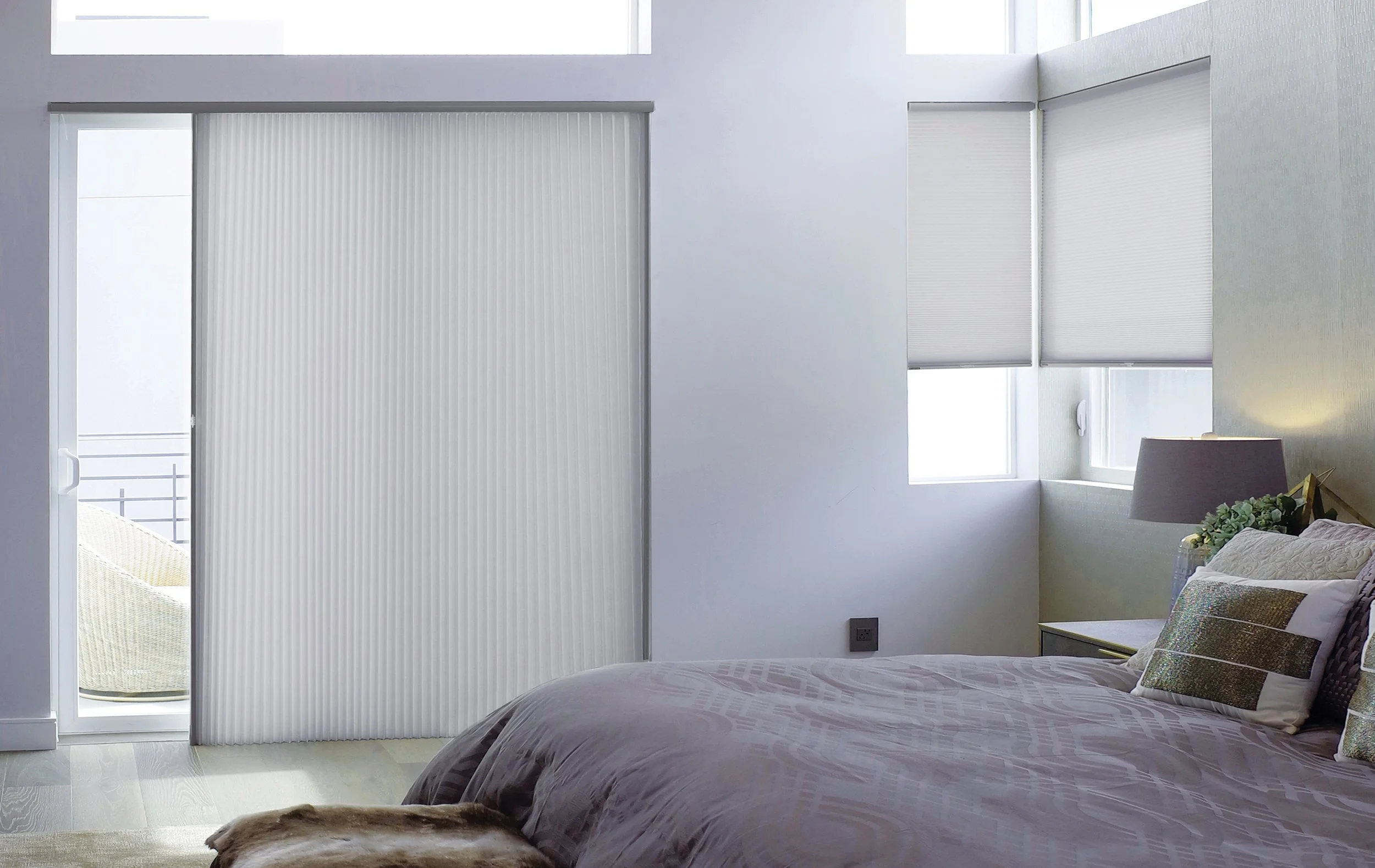 Modern bedroom with large sliding glass door, white blinds, partial view of balcony, and neatly made bed with pillows and a lamp.