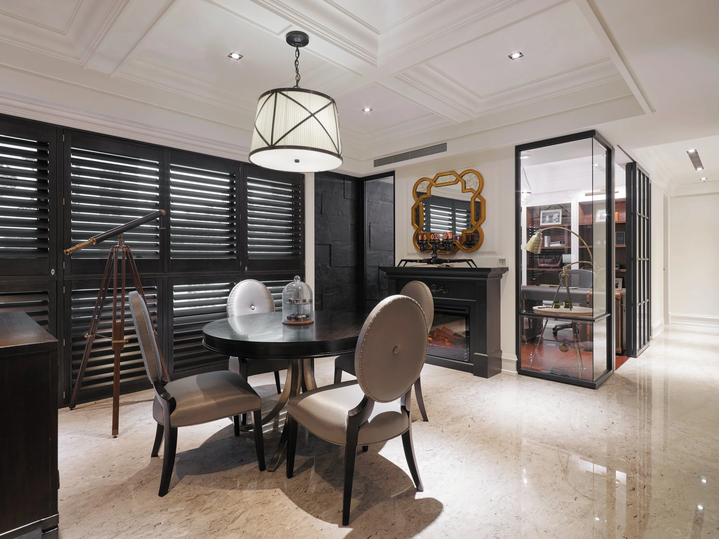 Modern dining room with black shuttered windows, a round dining table with four beige upholstered chairs, a decorative fireplace with a mirror above, and a glass-enclosed office space to the right, all with polished marble flooring.