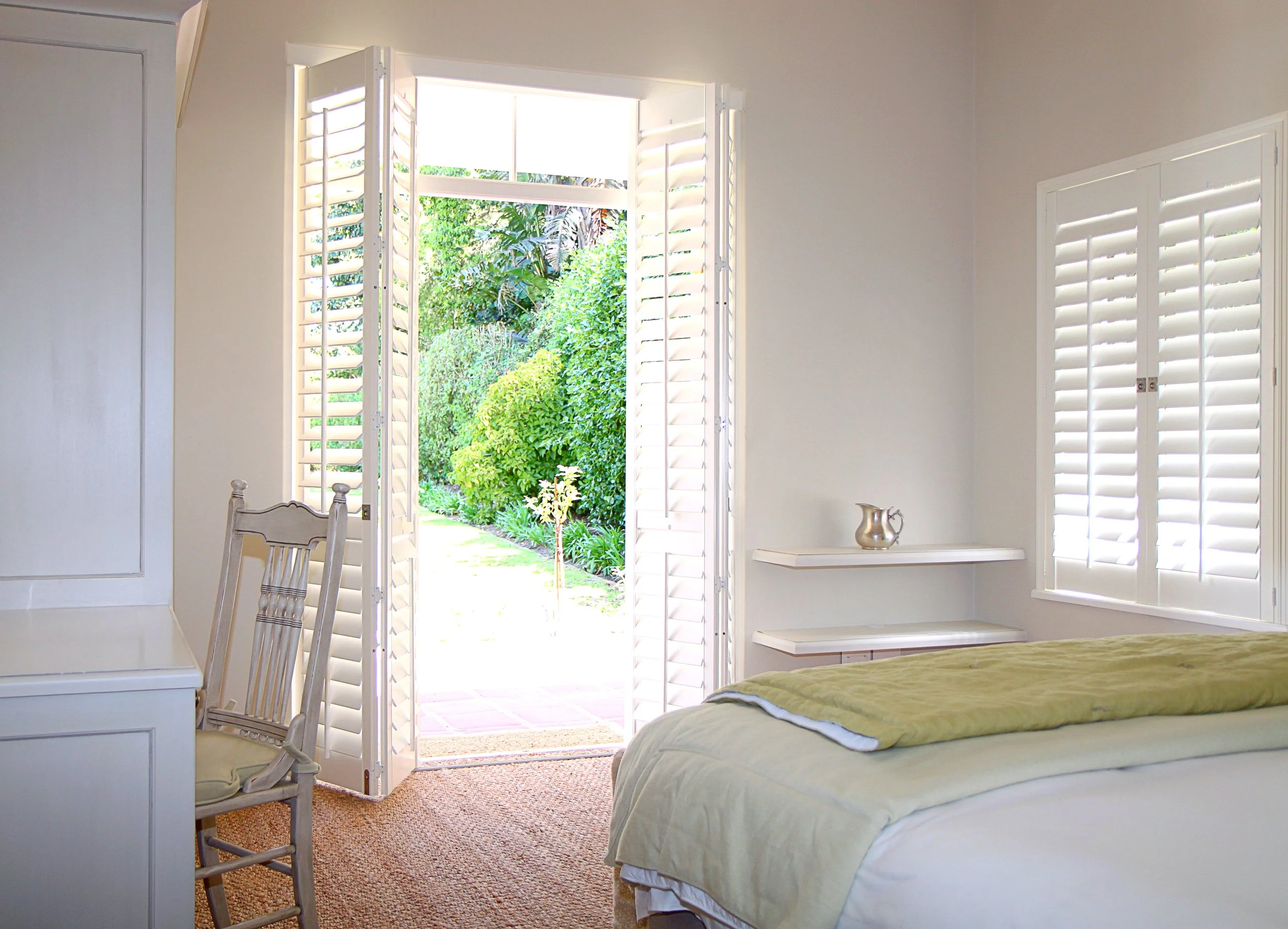 A bedroom with an open door leading to a lush green garden outside. The room has white walls, a bed with a pale yellow blanket, a silver pitcher on a white shelf, a white chair, and white window shutters.