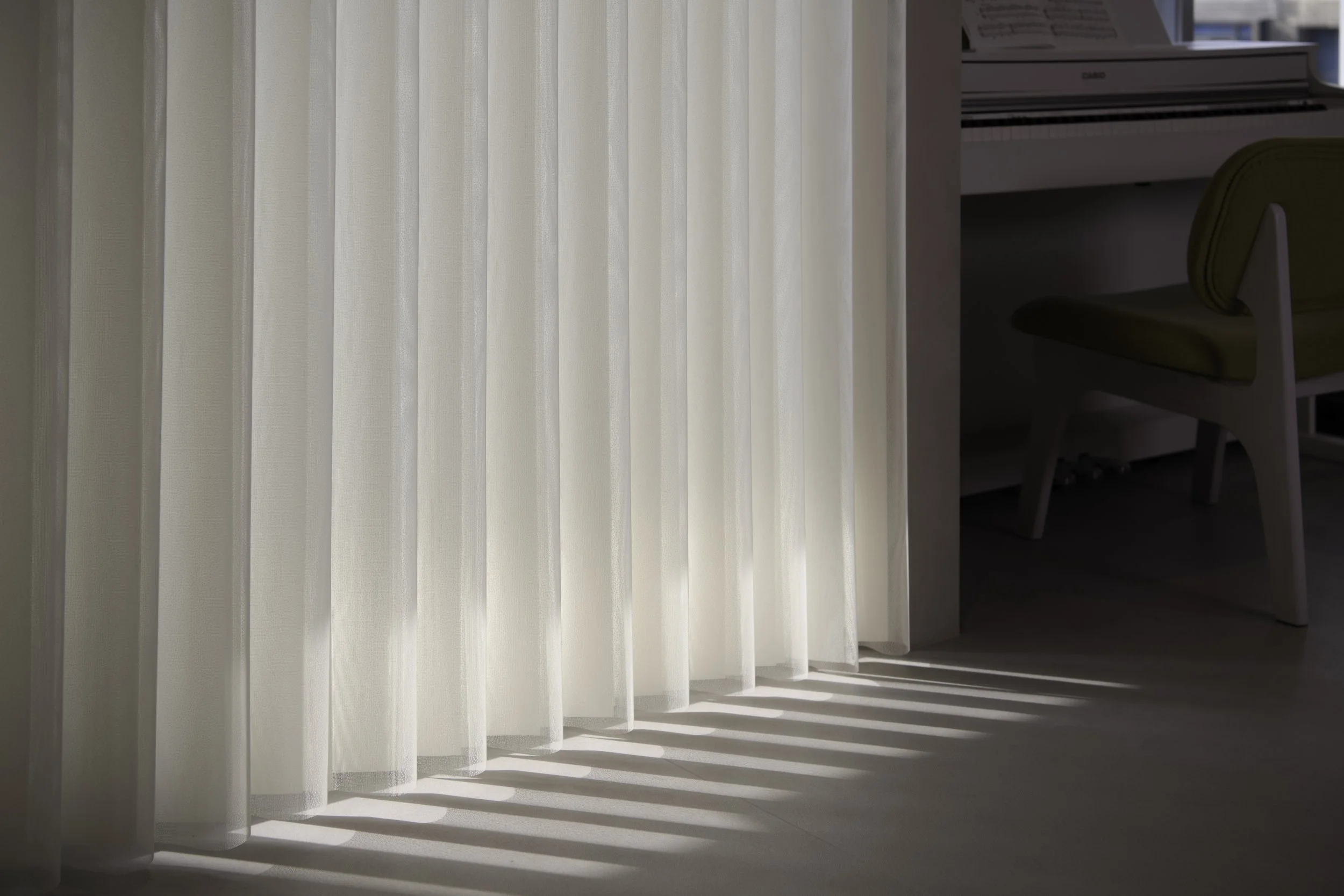 Sunlight filtering through white curtains, casting striped shadows on the floor in a room with a beige chair and a white desk.