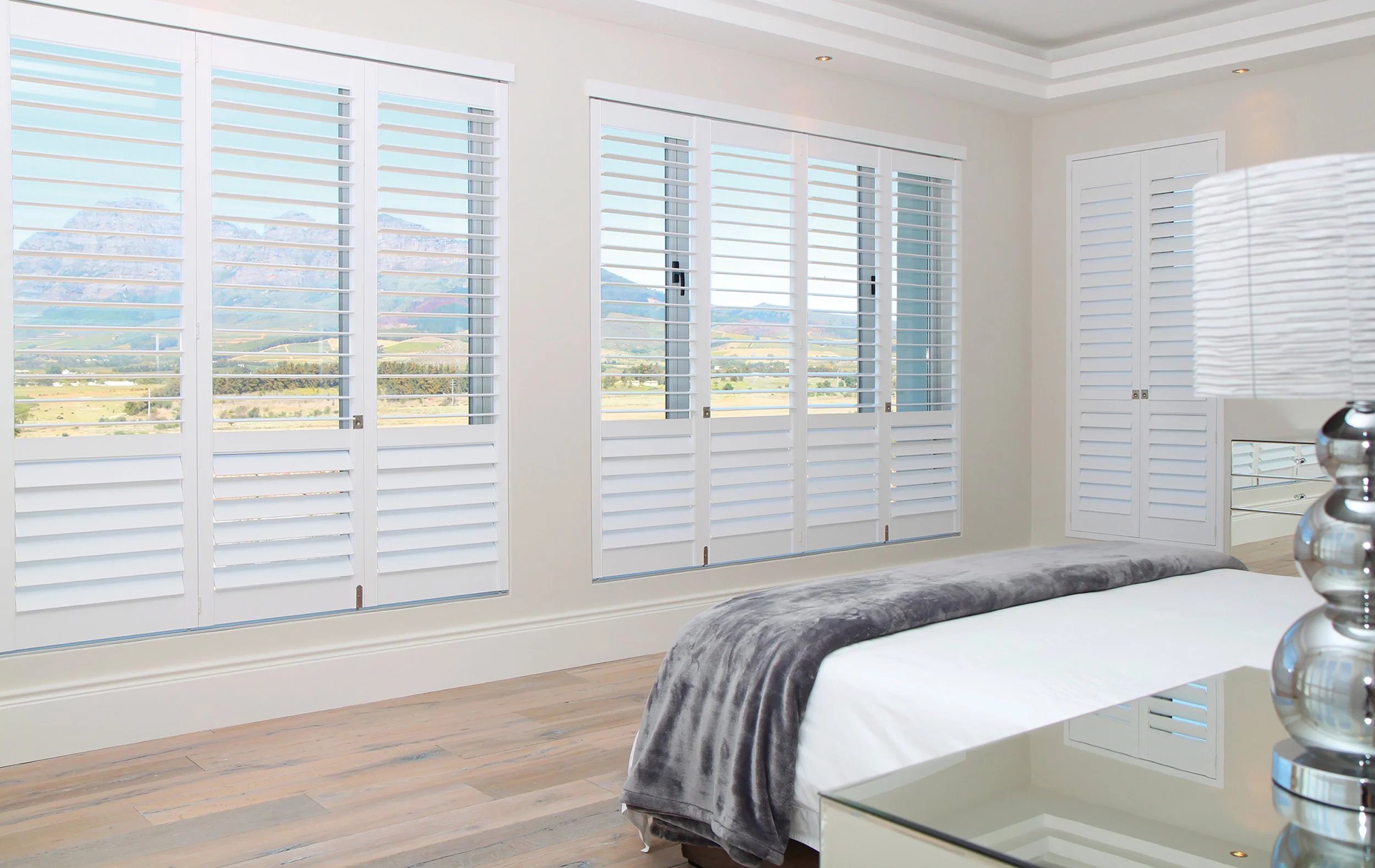 Bright bedroom with large windows and white plantation shutters, view of mountains and fields outside, bed with white bedding and gray throw, mirrored nightstand with silver lamp.