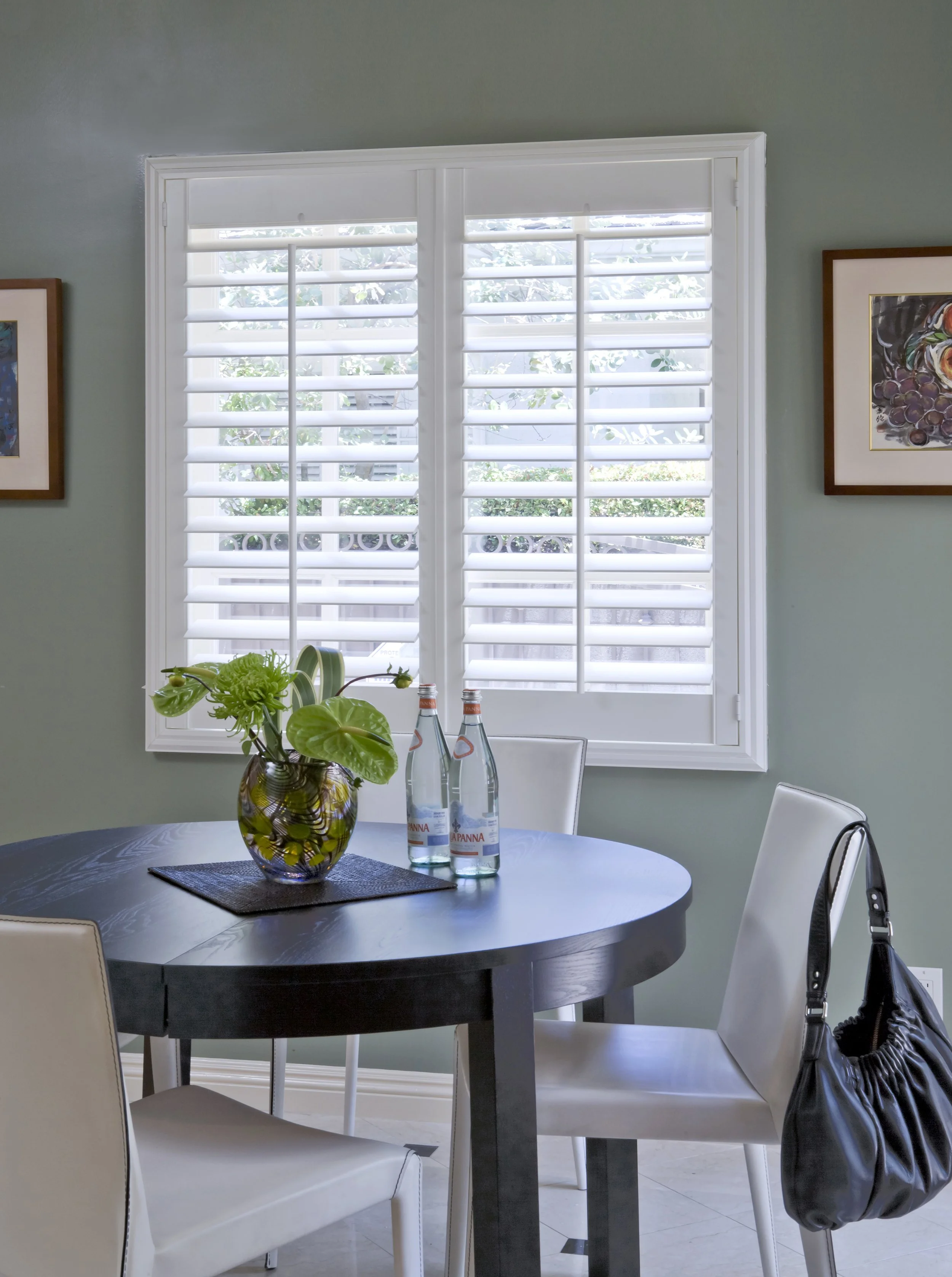 A dining room with a round black table, three white chairs, and a window with white shutters. On the table, there is a vase with green plants and two bottles of sparkling water. Two framed artworks hang on the green wall.