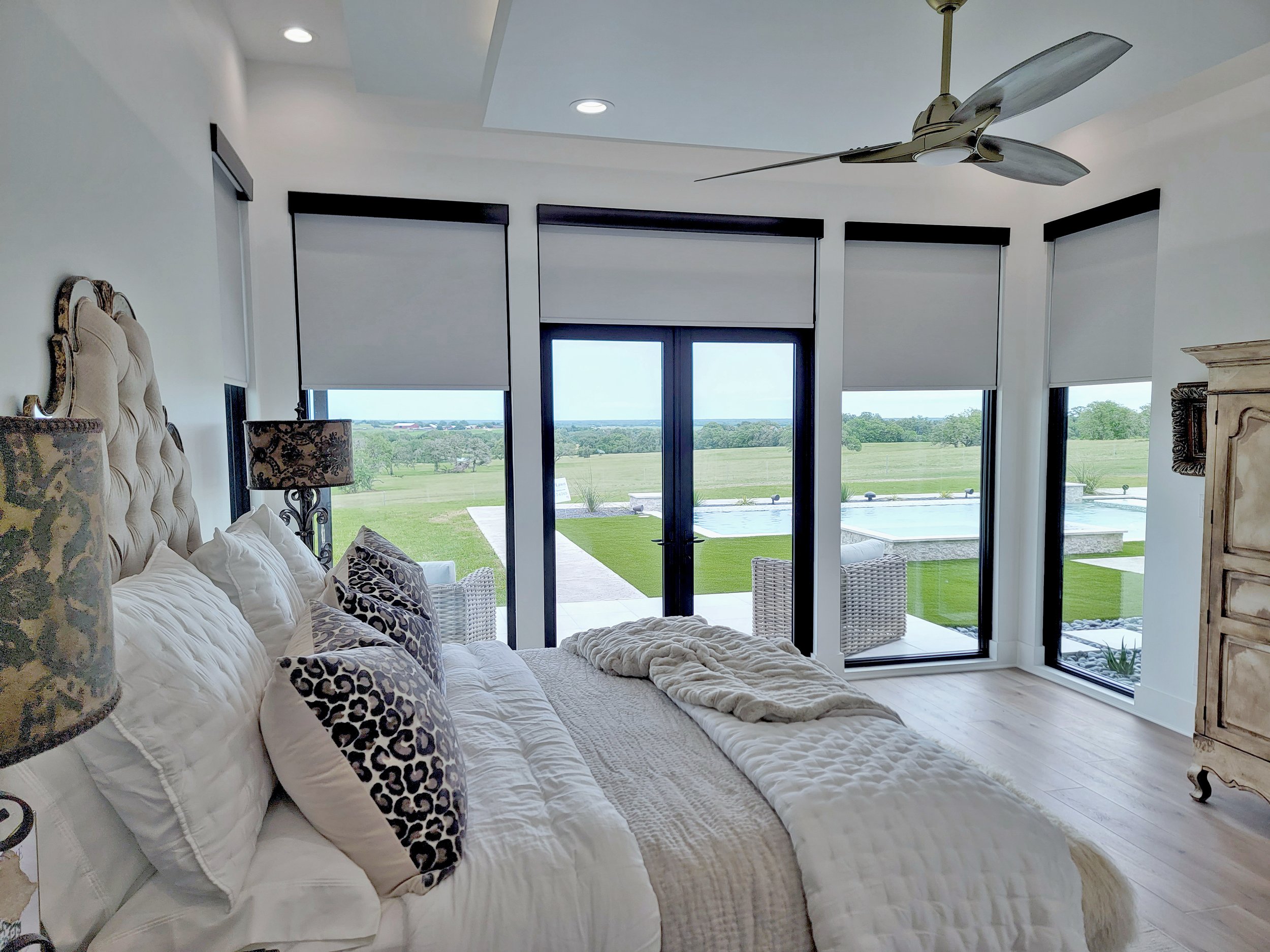 Bedroom with large windows overlooking a green lawn and pool, featuring a beige tufted headboard, white bedding, animal print pillows, side lamps, wicker furniture, and a ceiling fan.