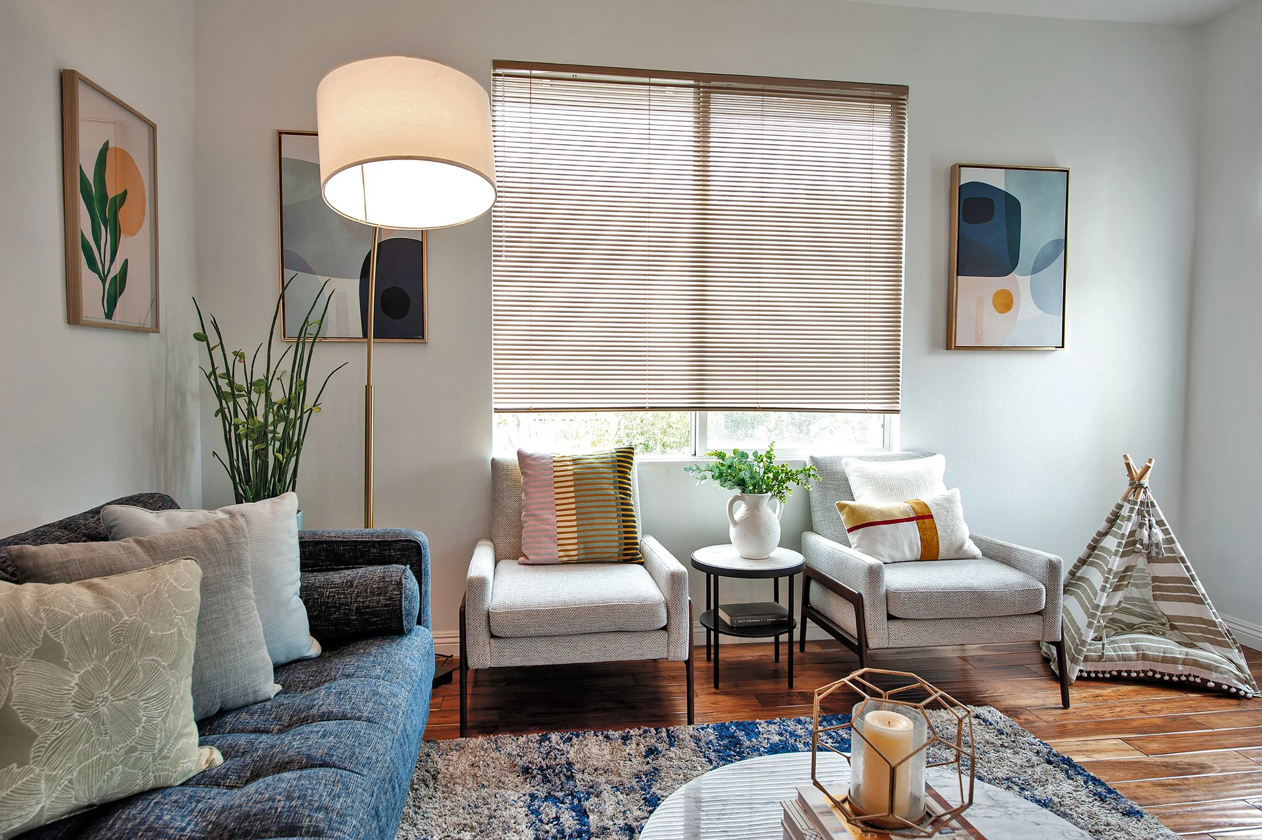 A living room with a gray couch, two white armchairs, a side table with a plant, a floor lamp, and art on the walls. There's a window with blinds, a colorful rug, and a small teepee in the corner.