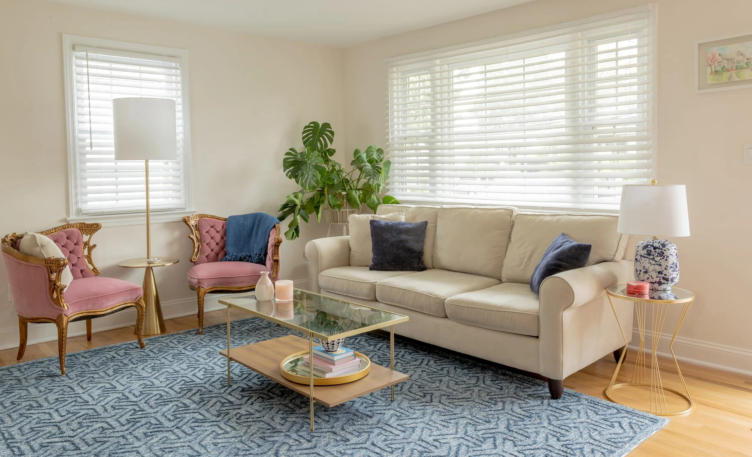 A cozy living room with a beige sofa, blue pillows, a glass-top coffee table with books and candles, pink Victorian chairs, a tall white floor lamp, a gold side table with a lamp, a large green plant, and white window blinds letting in natural light.