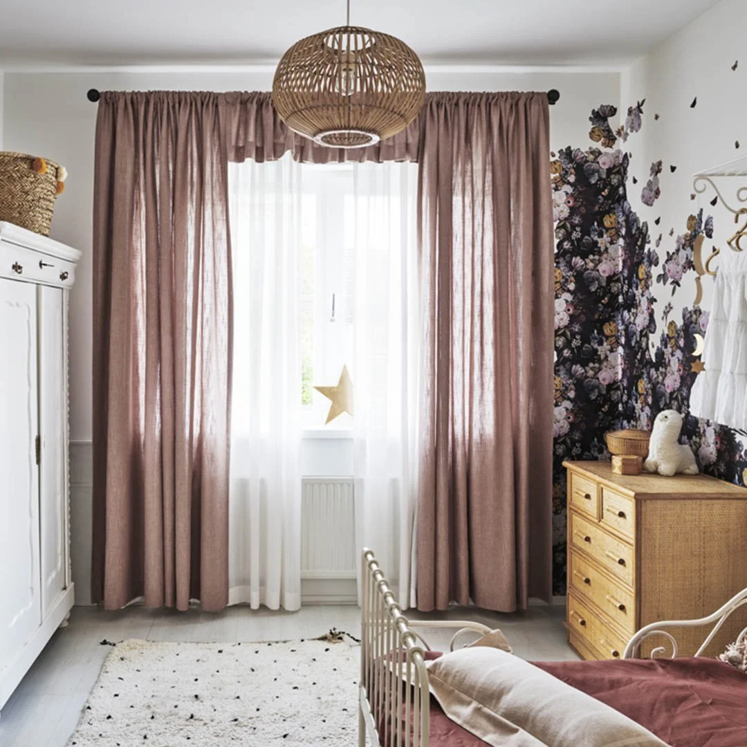 A bedroom with white walls and wooden flooring, featuring a window with sheer white and pink curtains, floral decor on the wall, a white wardrobe, a wooden dresser, and a bed with a soft pink blanket and stuffed animal.