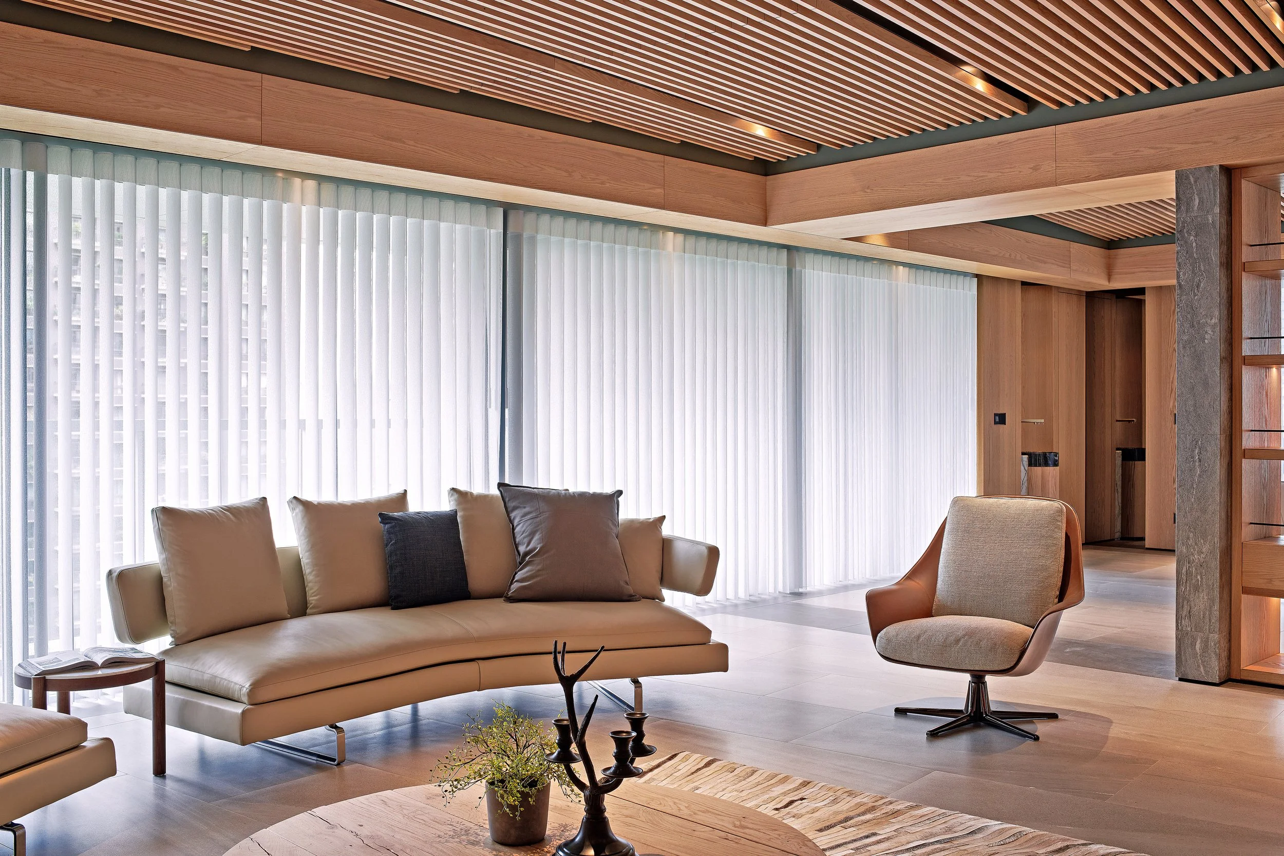 Modern living room with beige sofa, pillows, and a beige armchair in front of large windows with white vertical blinds, wooden ceiling with recessed lighting, and wooden wall accents.