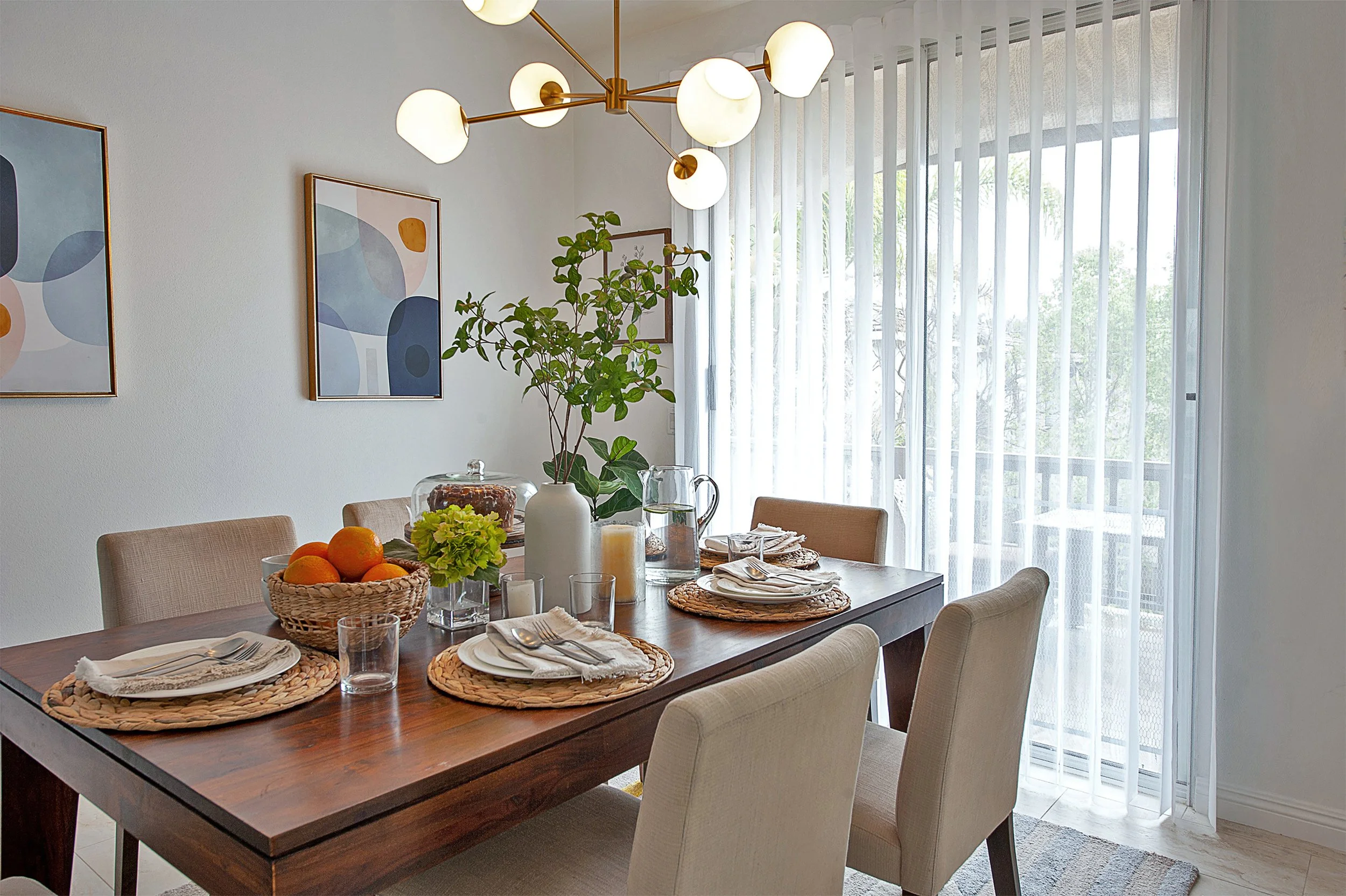 Dining room with a wooden table set for four, decorated with a basket of oranges, green plants, candles, and glassware. Modern art paintings hang on the wall, and sheer white curtains cover large sliding glass doors leading to an outdoor balcony.
