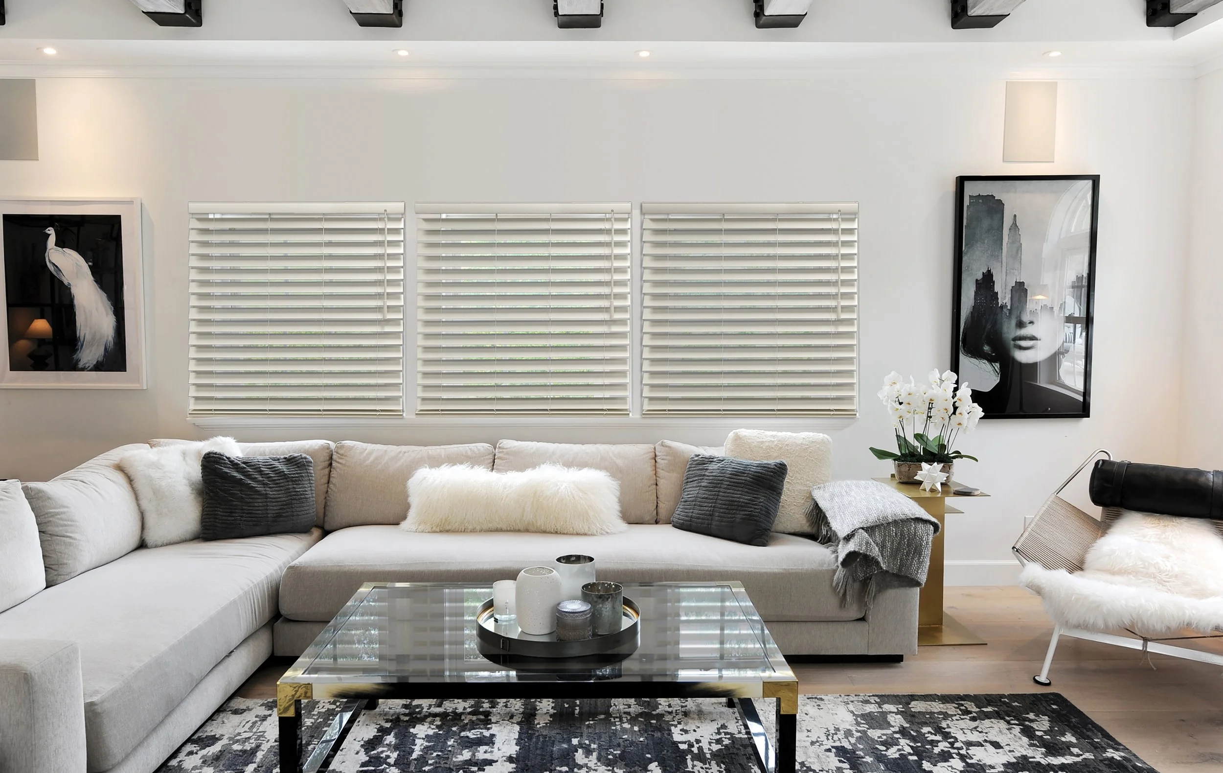 Modern living room with white sectional sofa, black and white artwork, white blinds, glass coffee table, and floral decor.