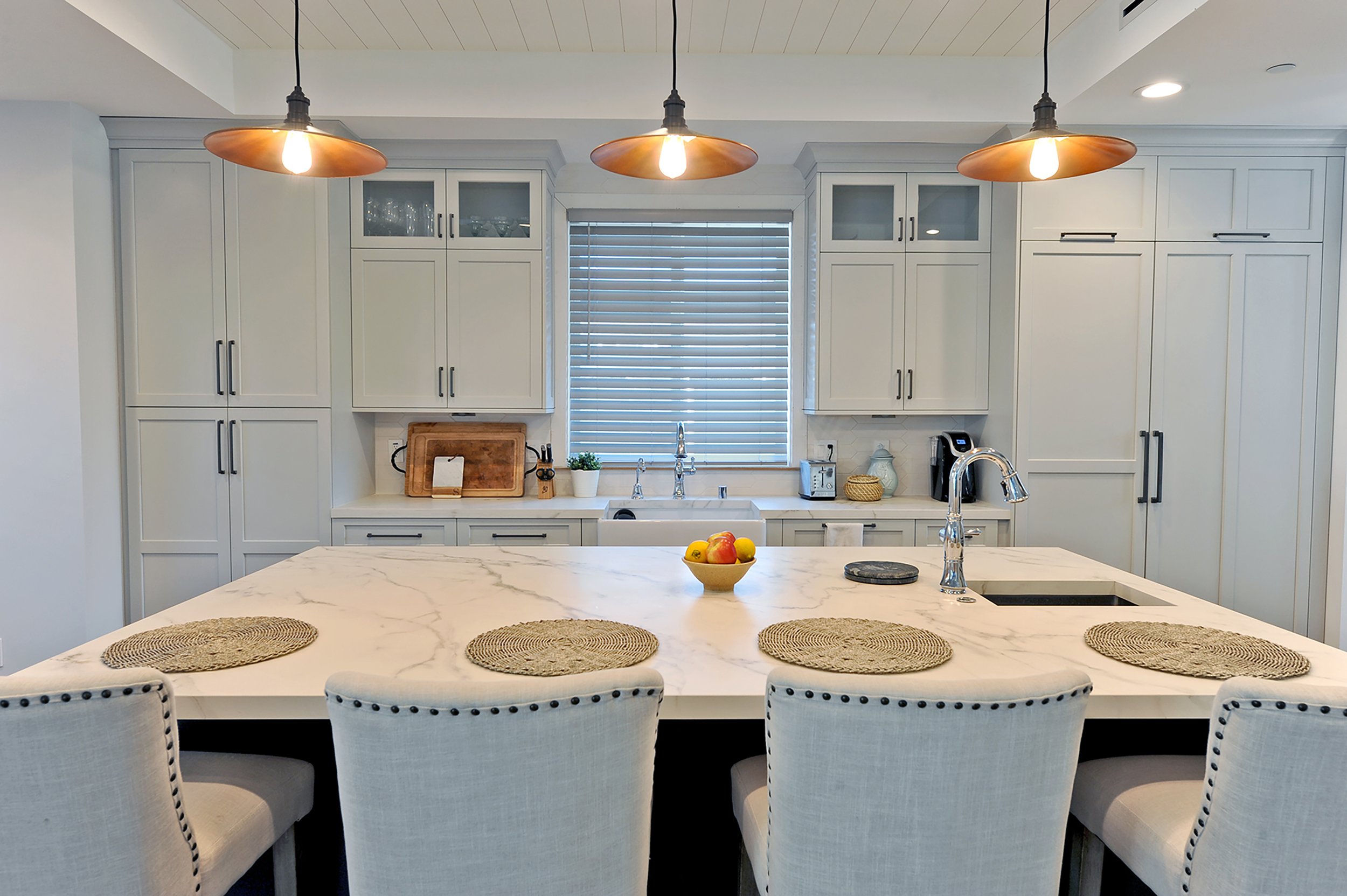 Modern white kitchen with marble island, four beige chairs, and cabinets, pendant lights, window with blinds, fruit bowl on island, and small appliances on counter.
